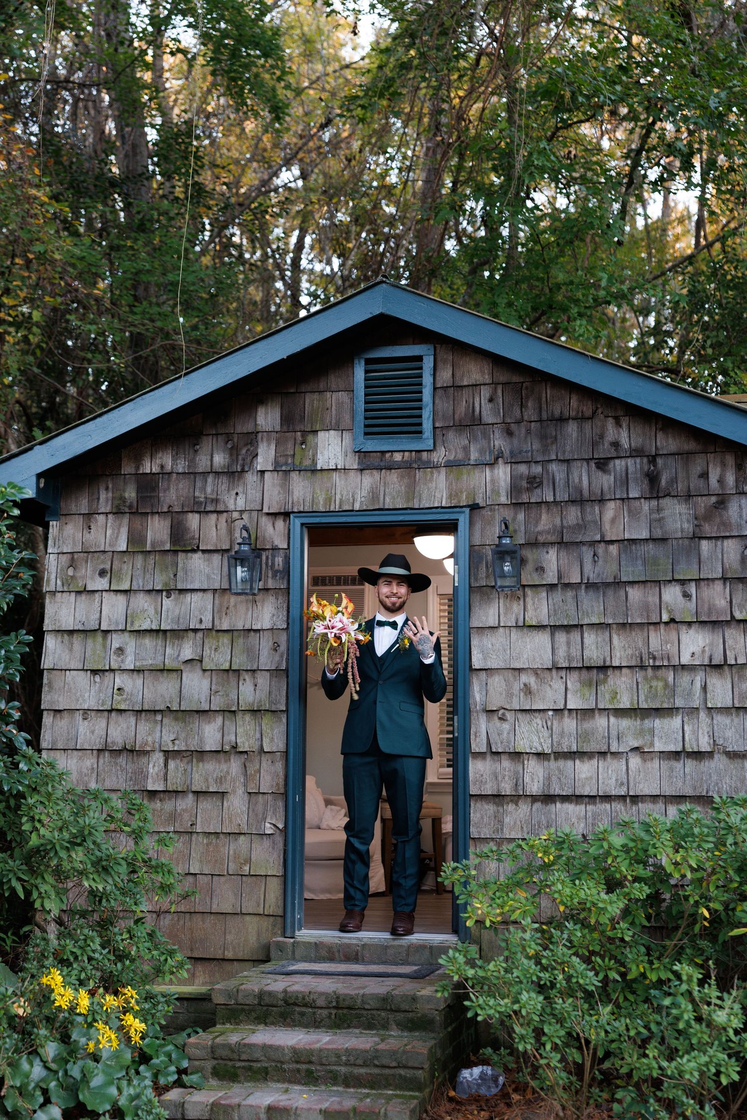 Groom in dark suit and cowboy hat stands in doorway of rustic cedar shingle cottage holding bouquet.