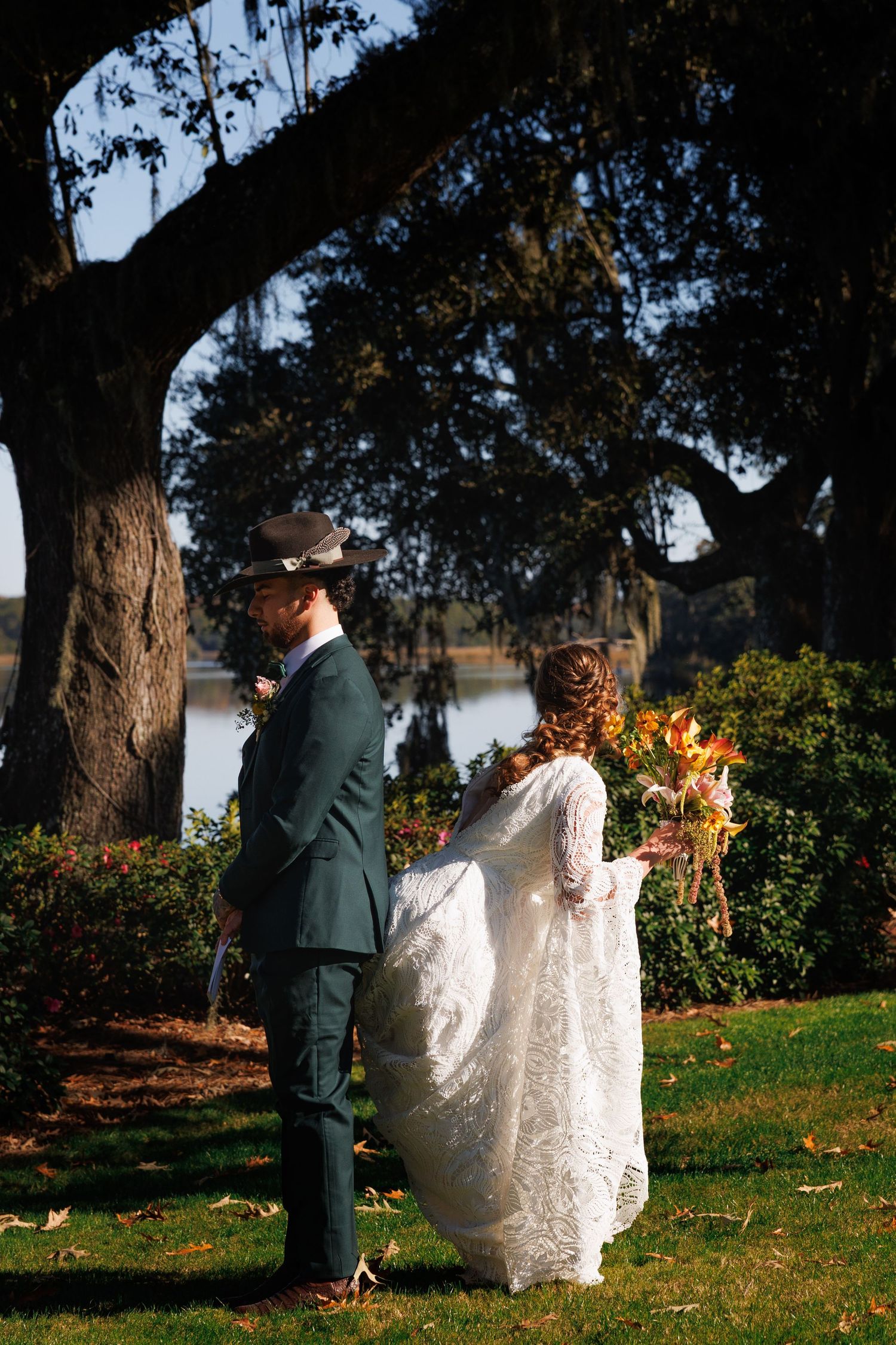 Bride in lace gown and groom in green suit share a first look under a moss-draped oak tree by a serene lake.