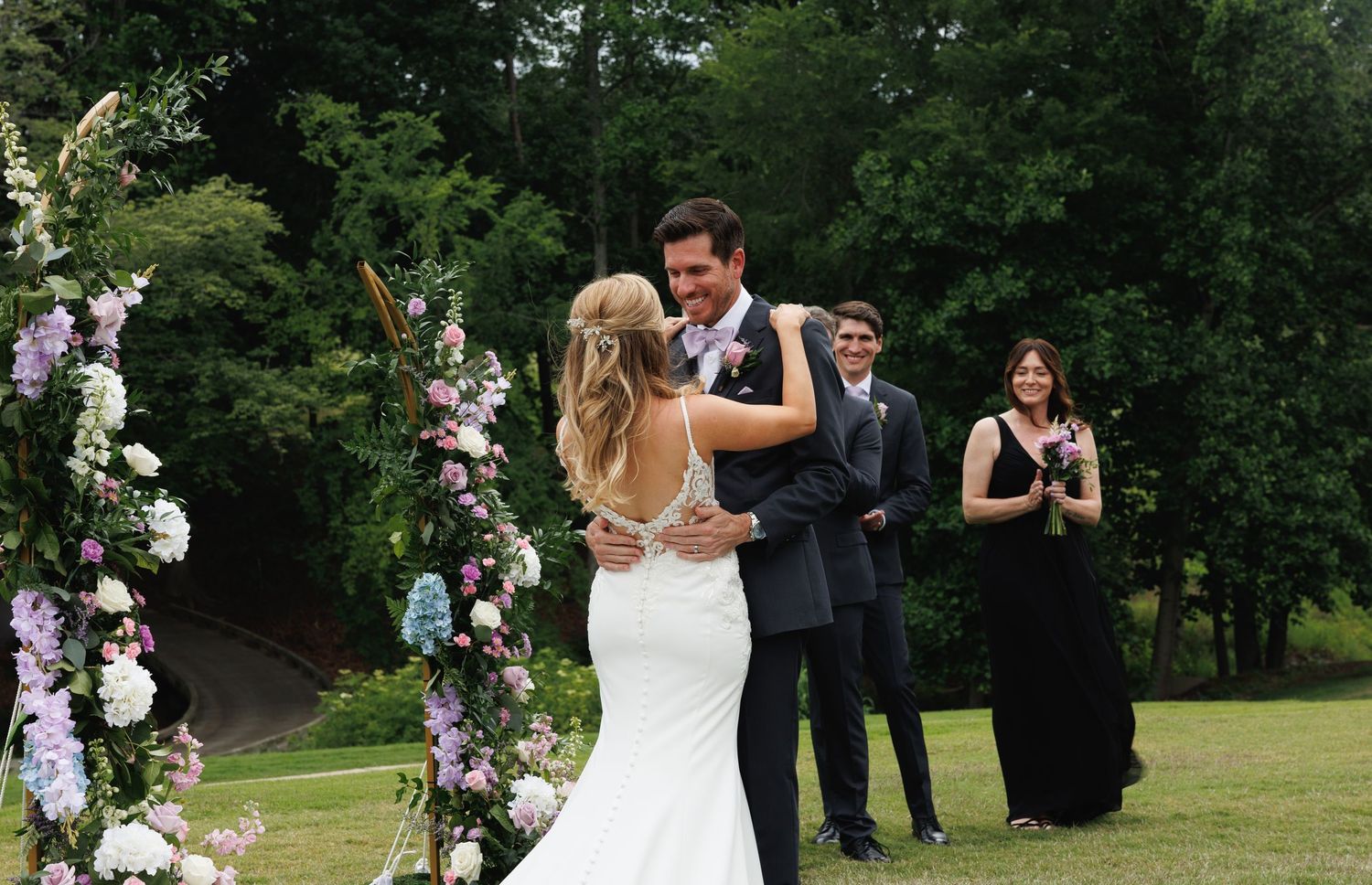 Bride and groom share first dance at outdoor wedding ceremony with floral arch and guests watching in background.