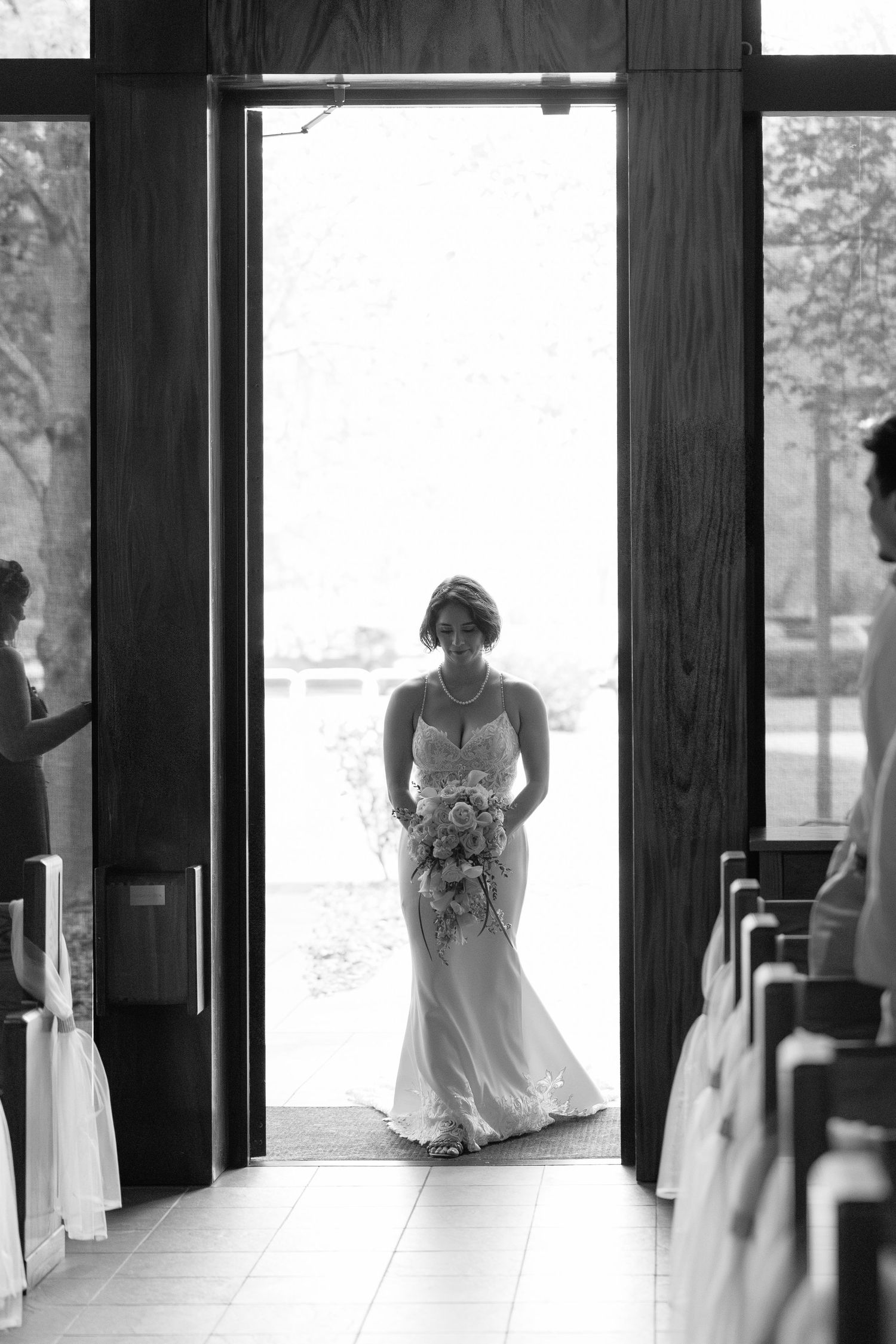 Bride in elegant white gown holds bouquet while standing in church doorway in black and white wedding photo.