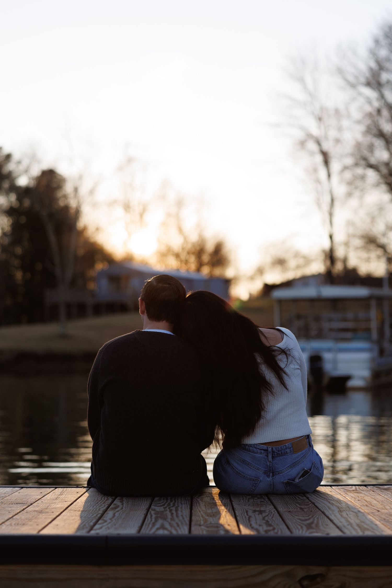 Romantic couple sitting on a wooden dock at sunset, woman resting head on mans shoulder.