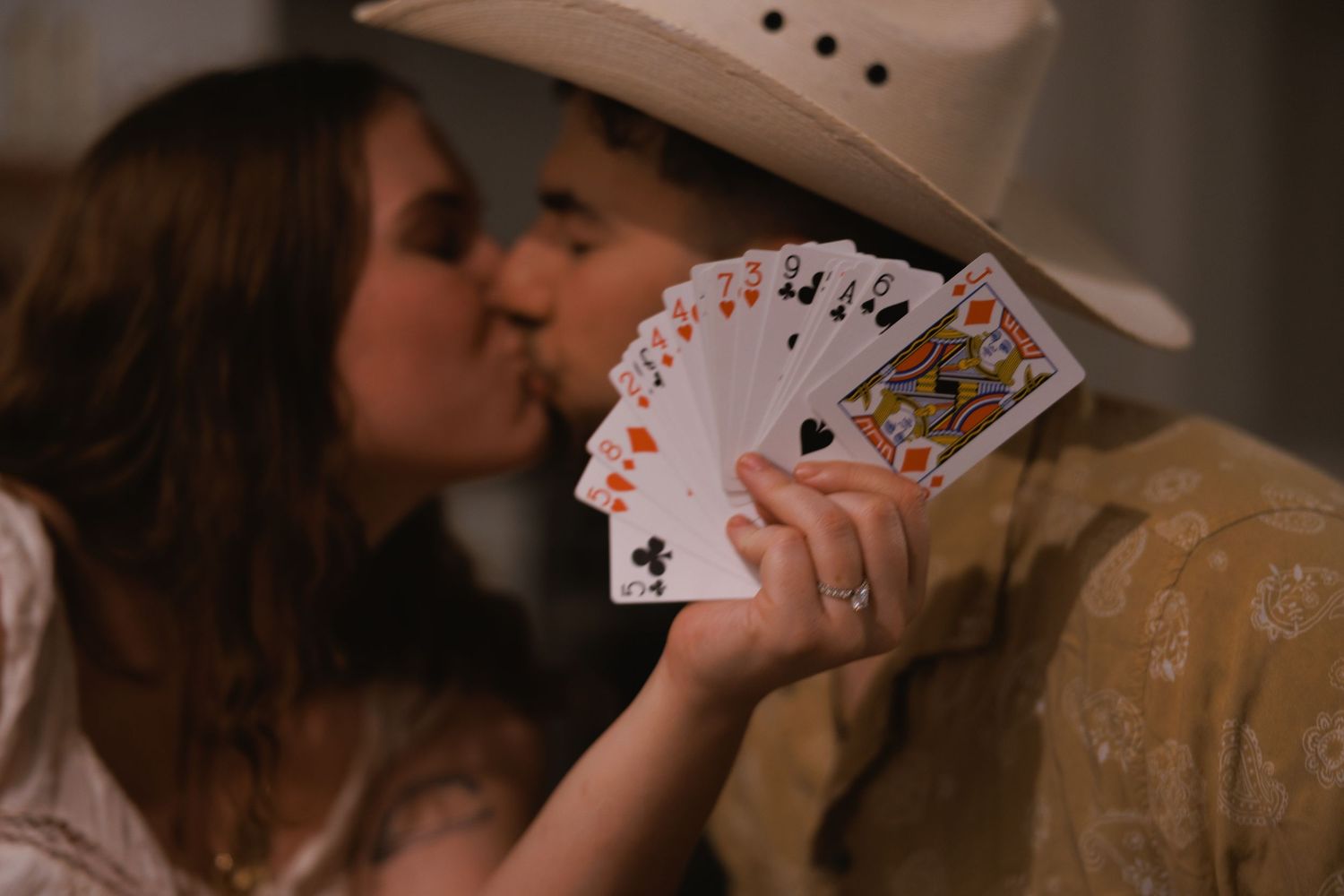 Couple sharing a romantic kiss behind a fan of playing cards, man wearing a white cowboy hat.