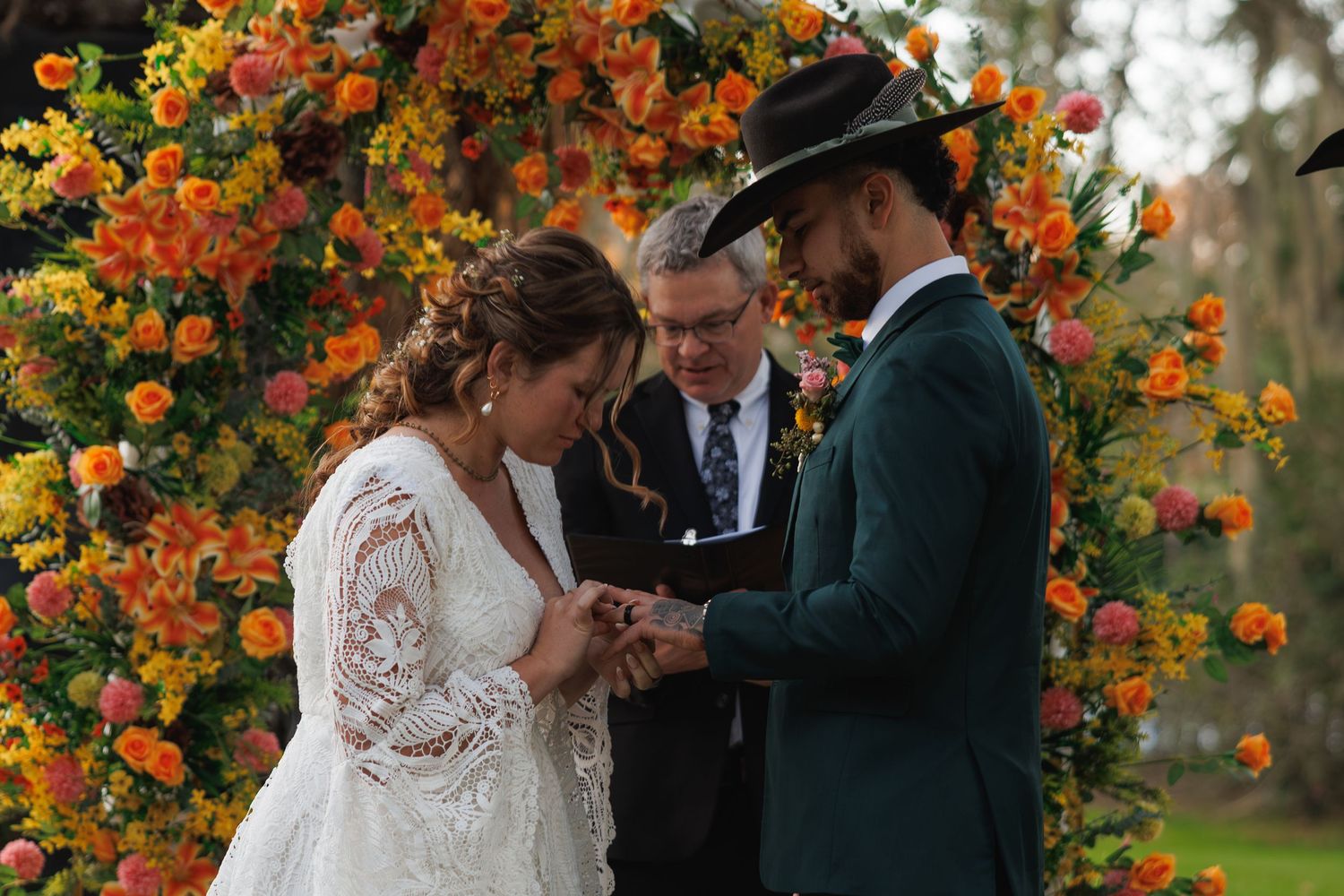 Bride and groom exchange rings at outdoor wedding ceremony beneath vibrant orange and yellow floral arch.