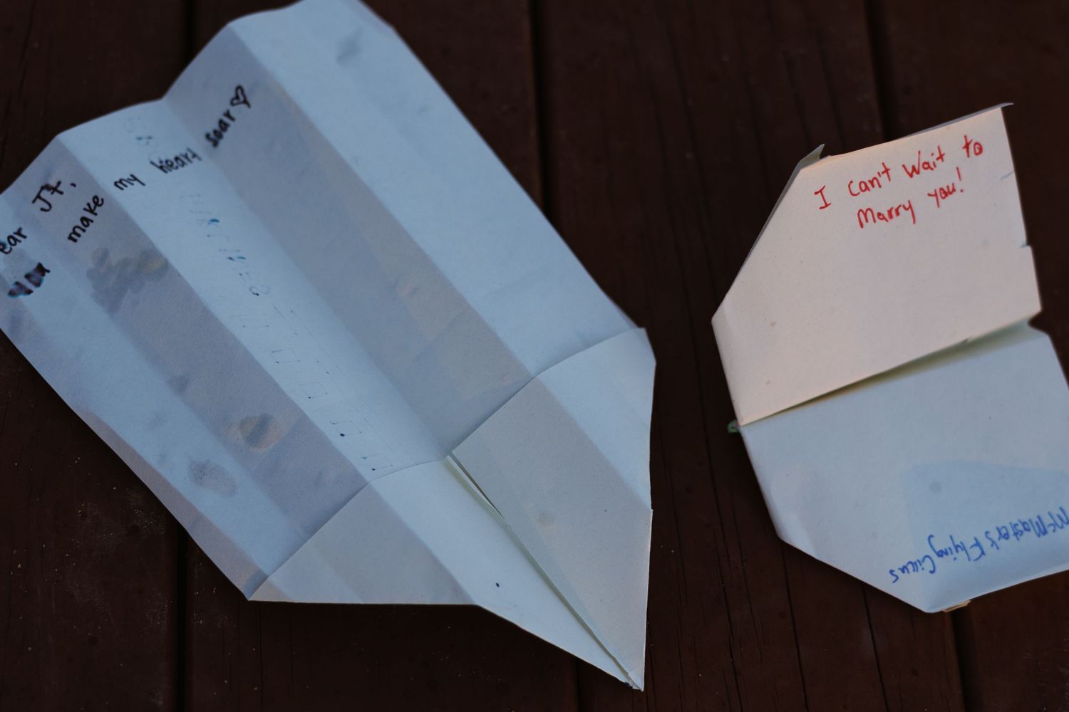 Two handwritten paper airplanes on a wooden surface with a marriage proposal message visible.