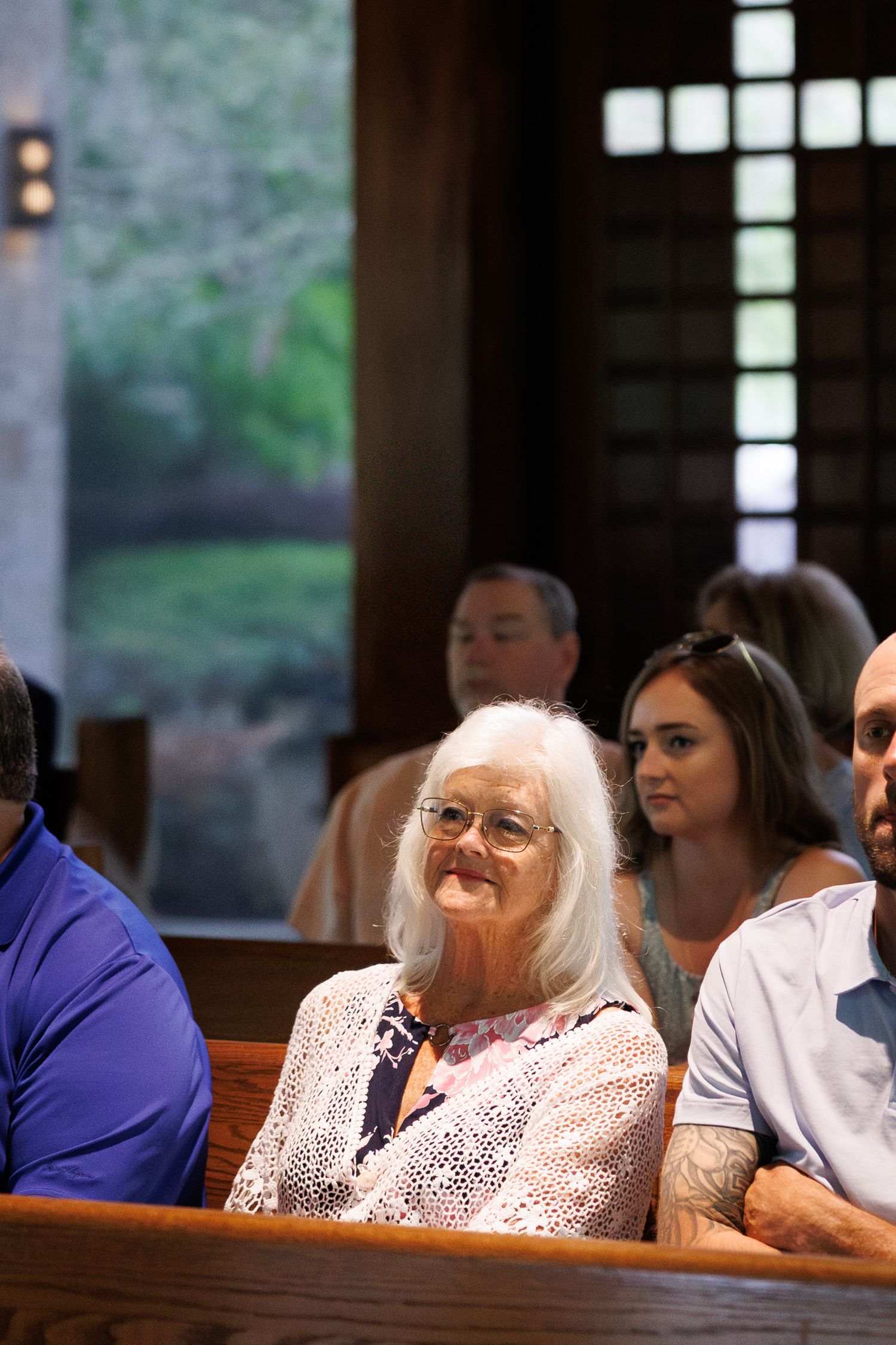 Elderly woman with white hair and glasses smiling warmly while seated in church wedding pew.