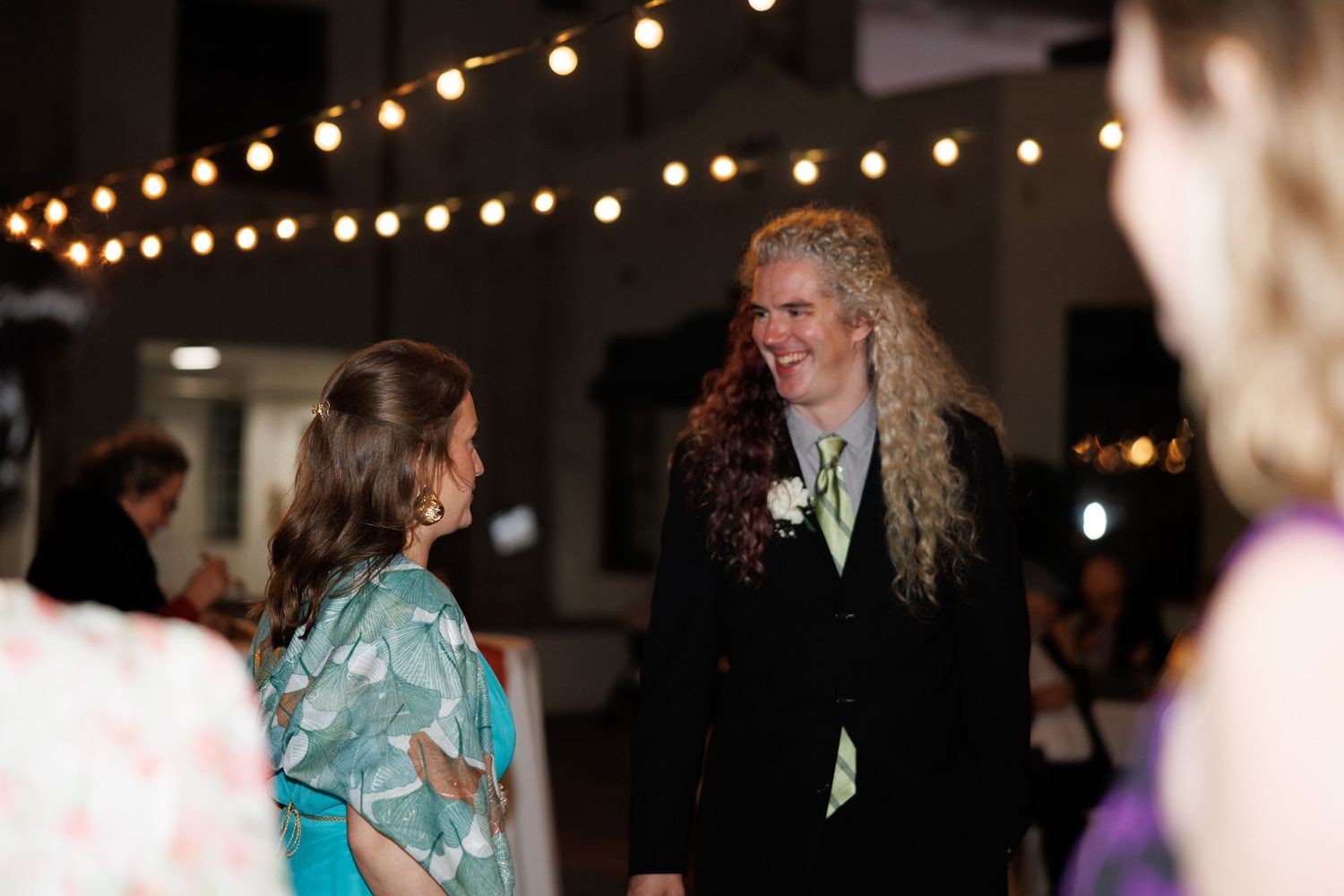 Man with long curly hair in a suit smiling and conversing with a woman at a wedding reception.