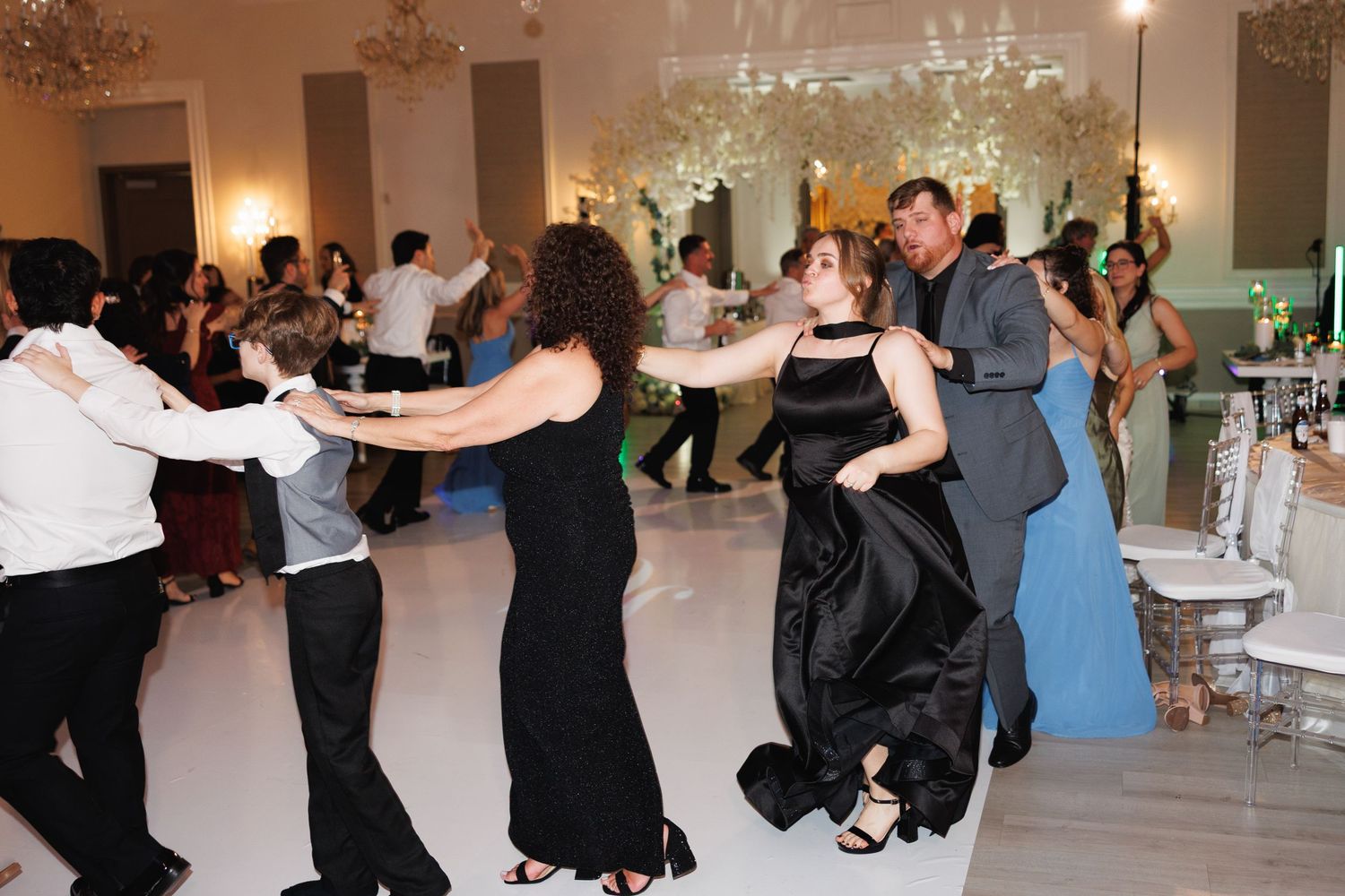 Wedding guests dancing energetically on a white dance floor beneath elegant floral decorations.