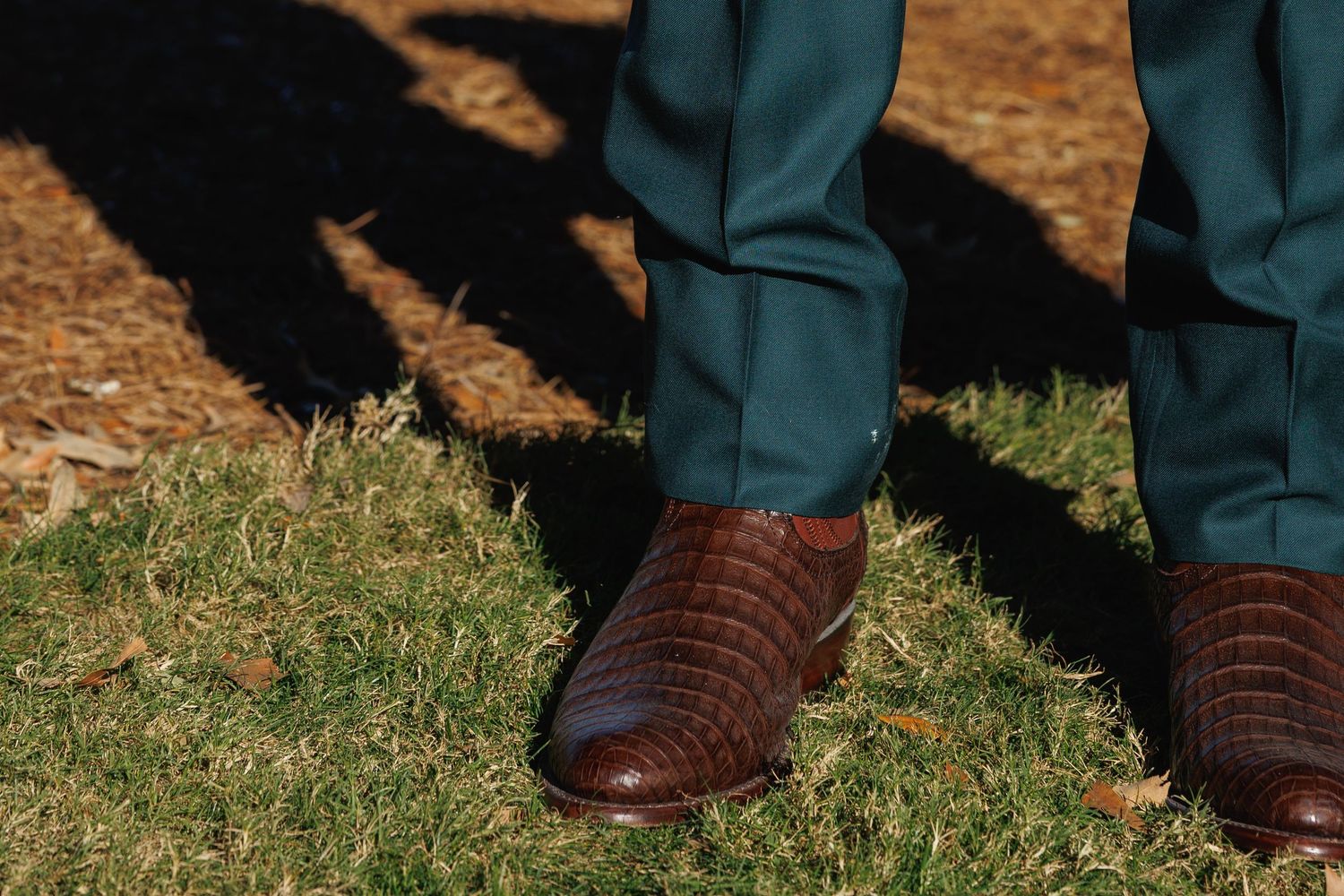 Man wearing teal dress pants and brown crocodile leather dress shoes standing on green grass outdoors.