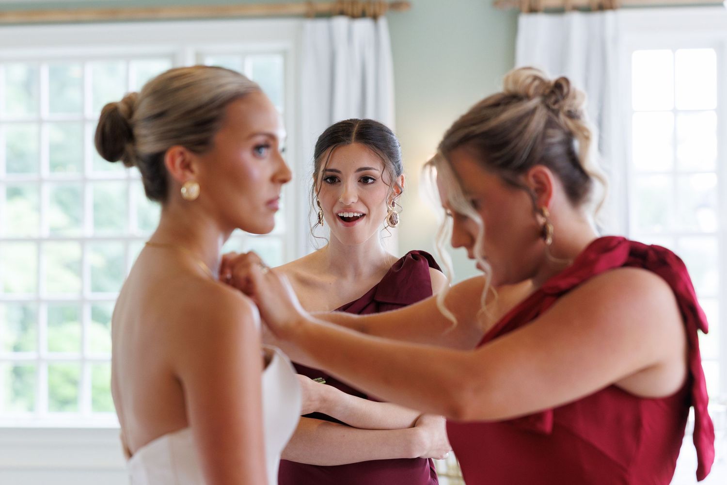 Bridesmaids in burgundy help bride adjust strapless wedding gown during wedding preparations.