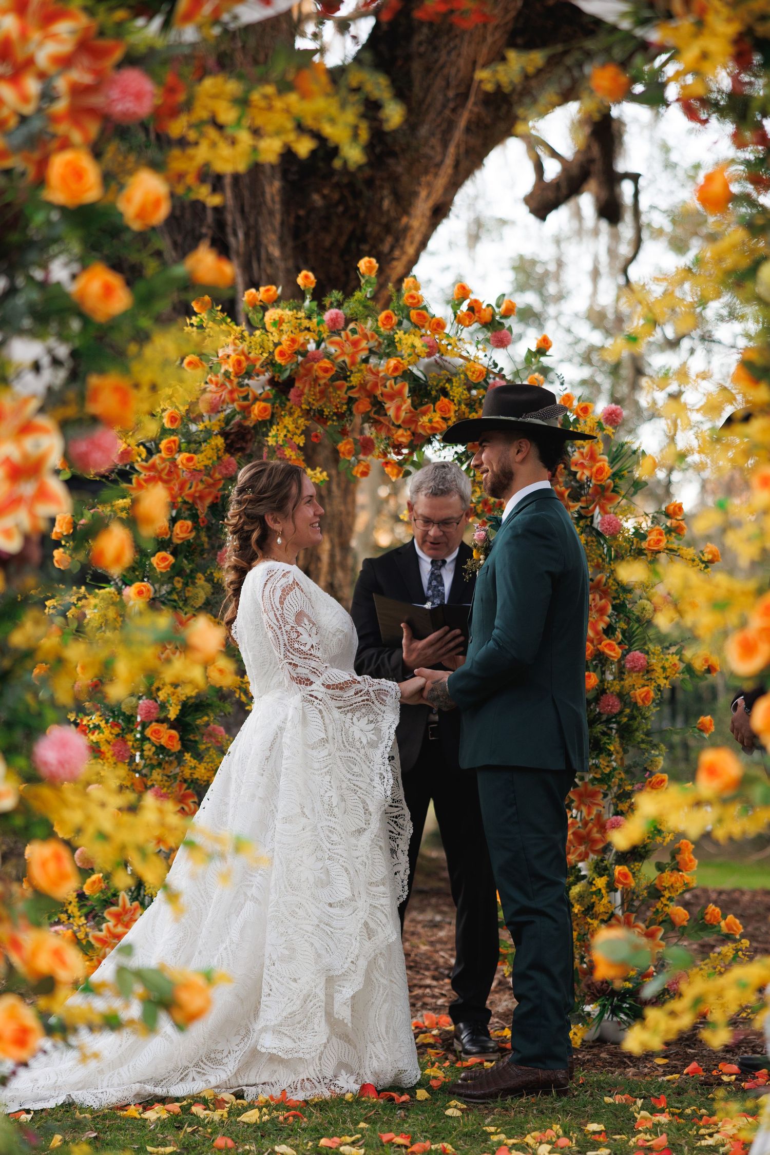 Bride and groom exchange vows under stunning orange and yellow floral arch at outdoor fall wedding ceremony.