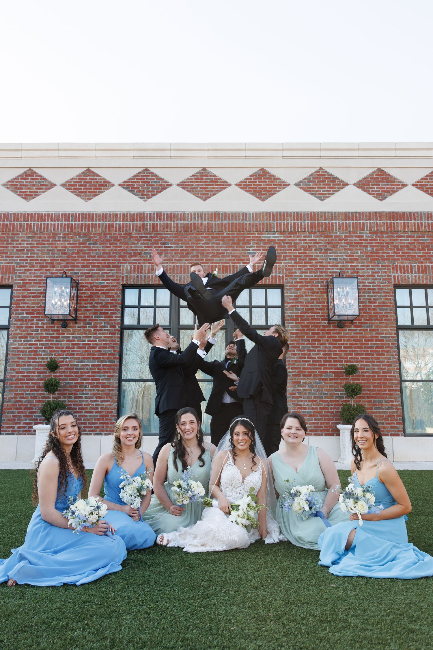 Wedding party poses outside brick venue, bridesmaids in blue and green gowns seated while groomsmen toss groom in air at Yorkmont Courtyard.