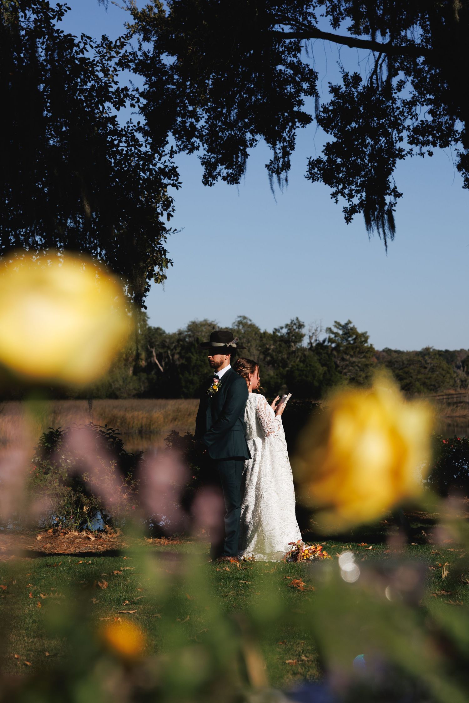 A bride and groom share a romantic moment outdoors, framed by yellow roses and Spanish moss-draped trees on a sunny day.