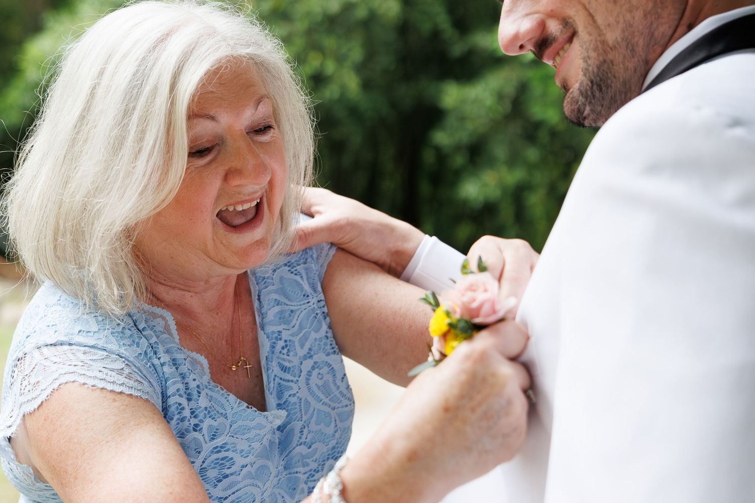 Elderly woman smiling while pinning a yellow boutonniere on a groom's white suit jacket at a wedding.
