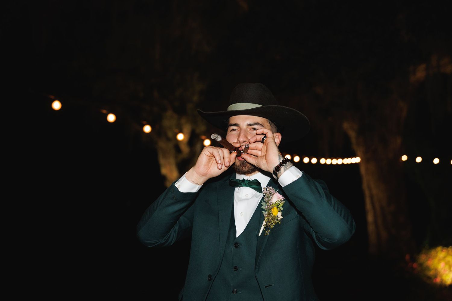 Groom in green suit bites cigar under string lights wearing a black cowboy hat at night.