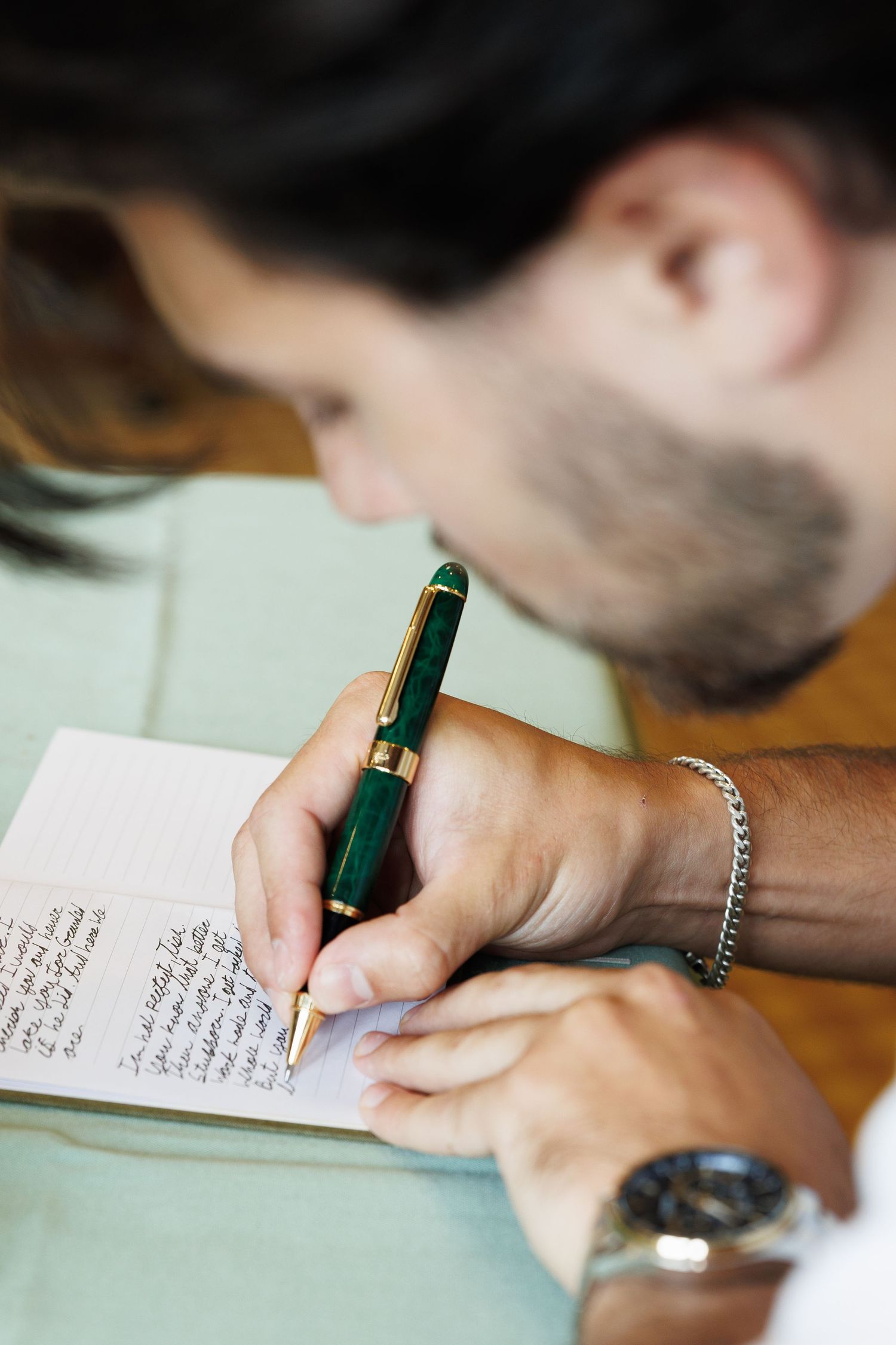 Groom writing heartfelt wedding vows in a small notebook with a green and gold fountain pen.