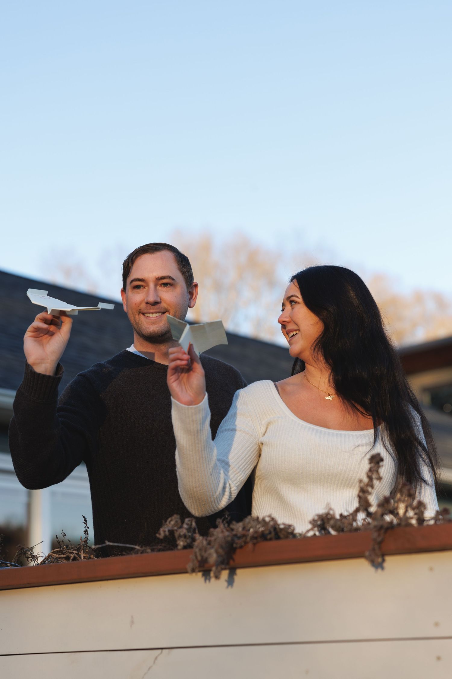 Smiling couple holding a paper airplane outdoors on a balcony with a clear blue sky background.