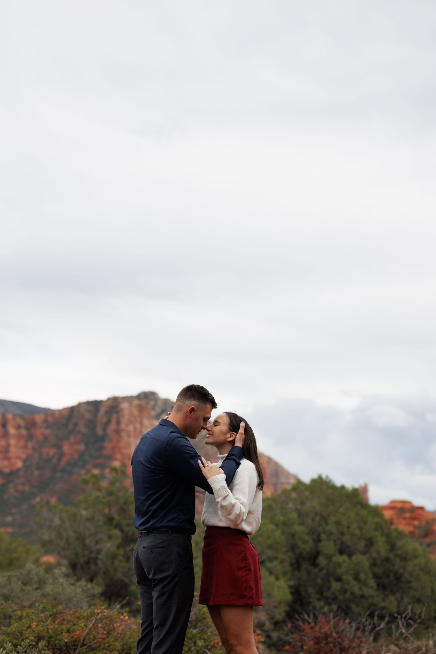 Engaged couple sharing a kiss amid Sedonas stunning red rock formations under cloudy skies.