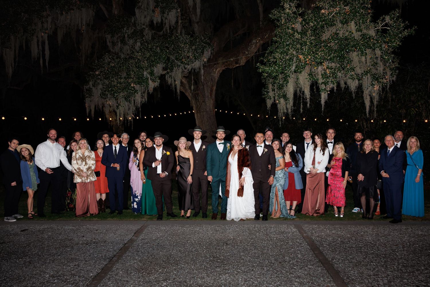 Large wedding party group photo outdoors at night under a tree with string lights at a formal celebration.