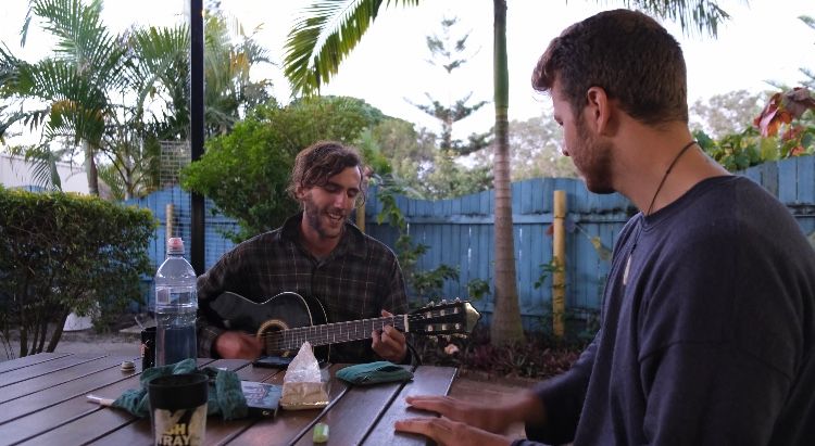 Two men relaxing outdoors, one playing acoustic guitar at a wooden table with drinks in a lush garden setting.