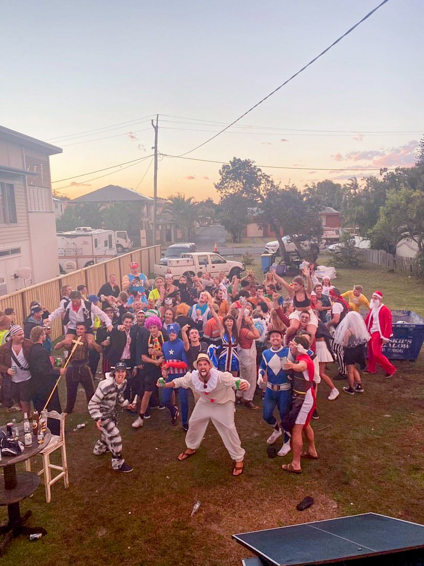 Large group of people in colorful costumes posing outdoors at a backyard costume party during sunset.