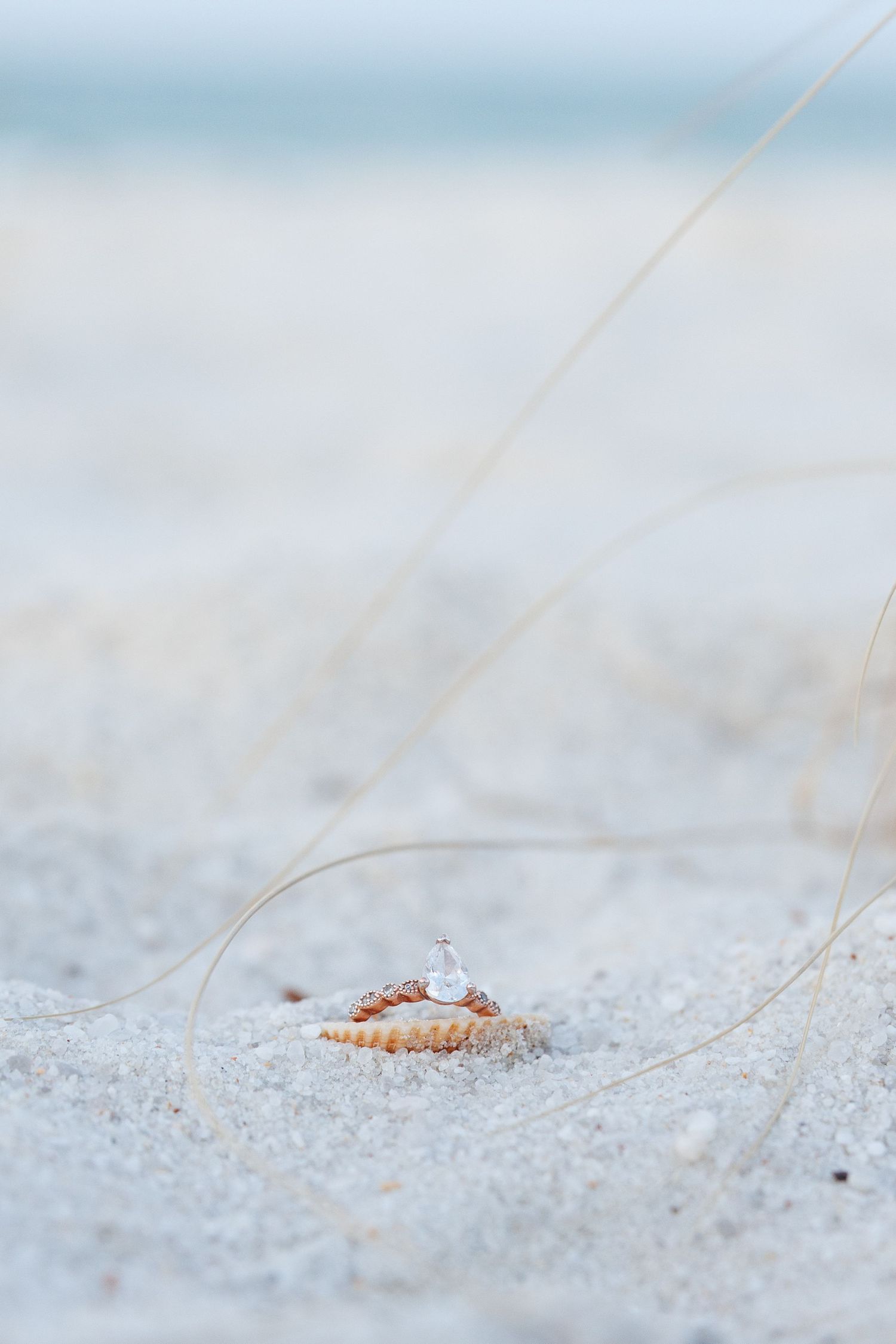 A diamond ring resting on a seashell in white sand on a beach with soft ocean waves in the background.