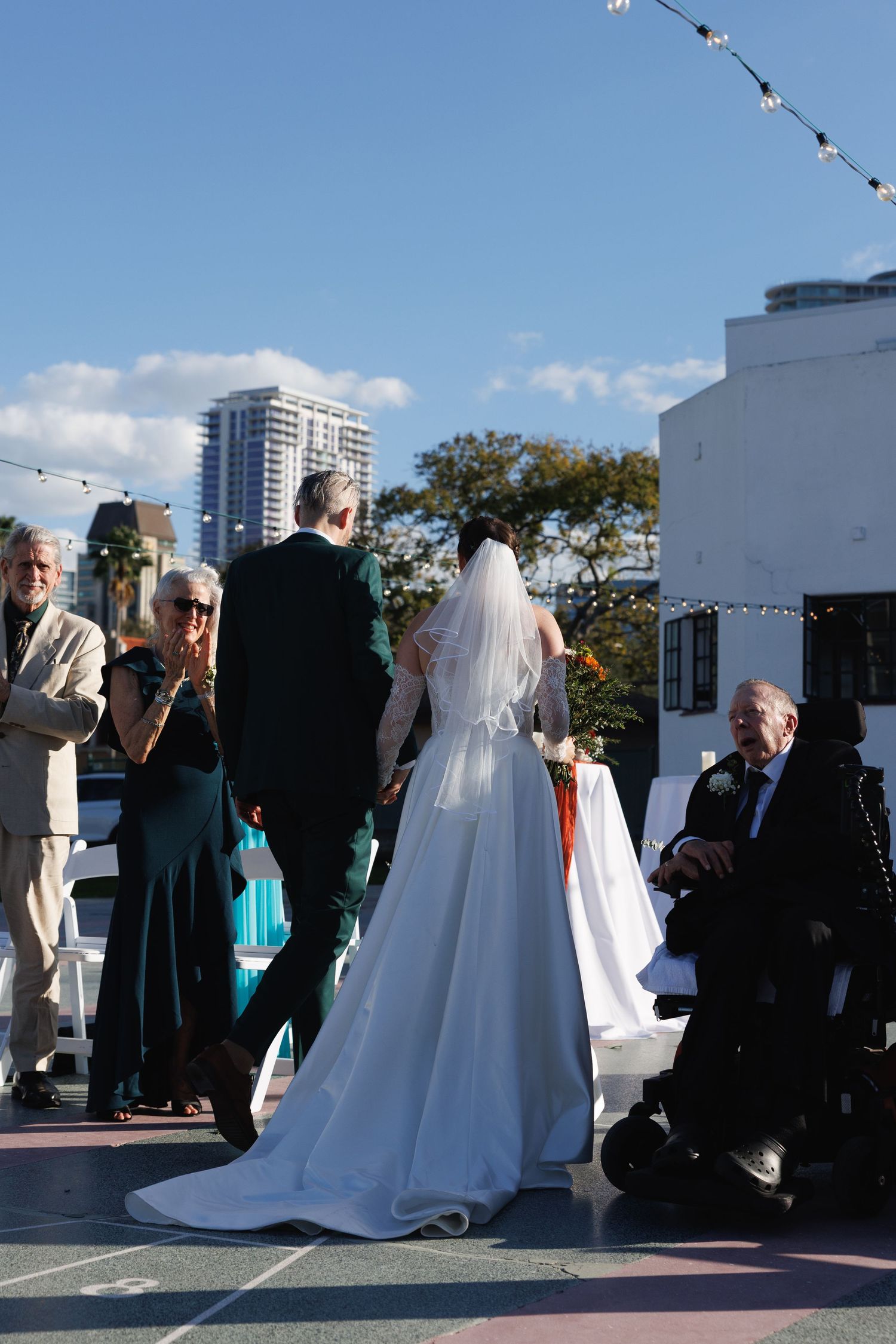 Bride in white gown and veil walks down outdoor rooftop aisle with guests and city skyline backdrop.
