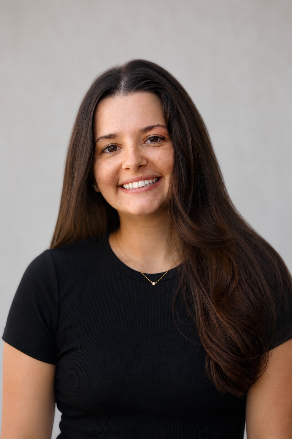 Smiling young woman with long brown hair wearing a black t-shirt poses for a professional headshot photo.