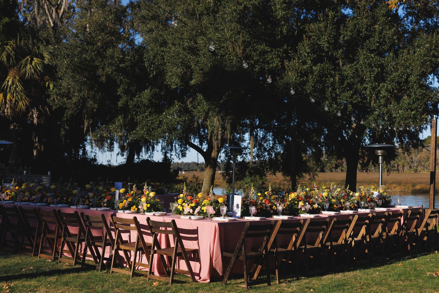 Outdoor wedding reception table with pink linens, yellow floral centerpieces, and wooden chairs set near a scenic waterfront.