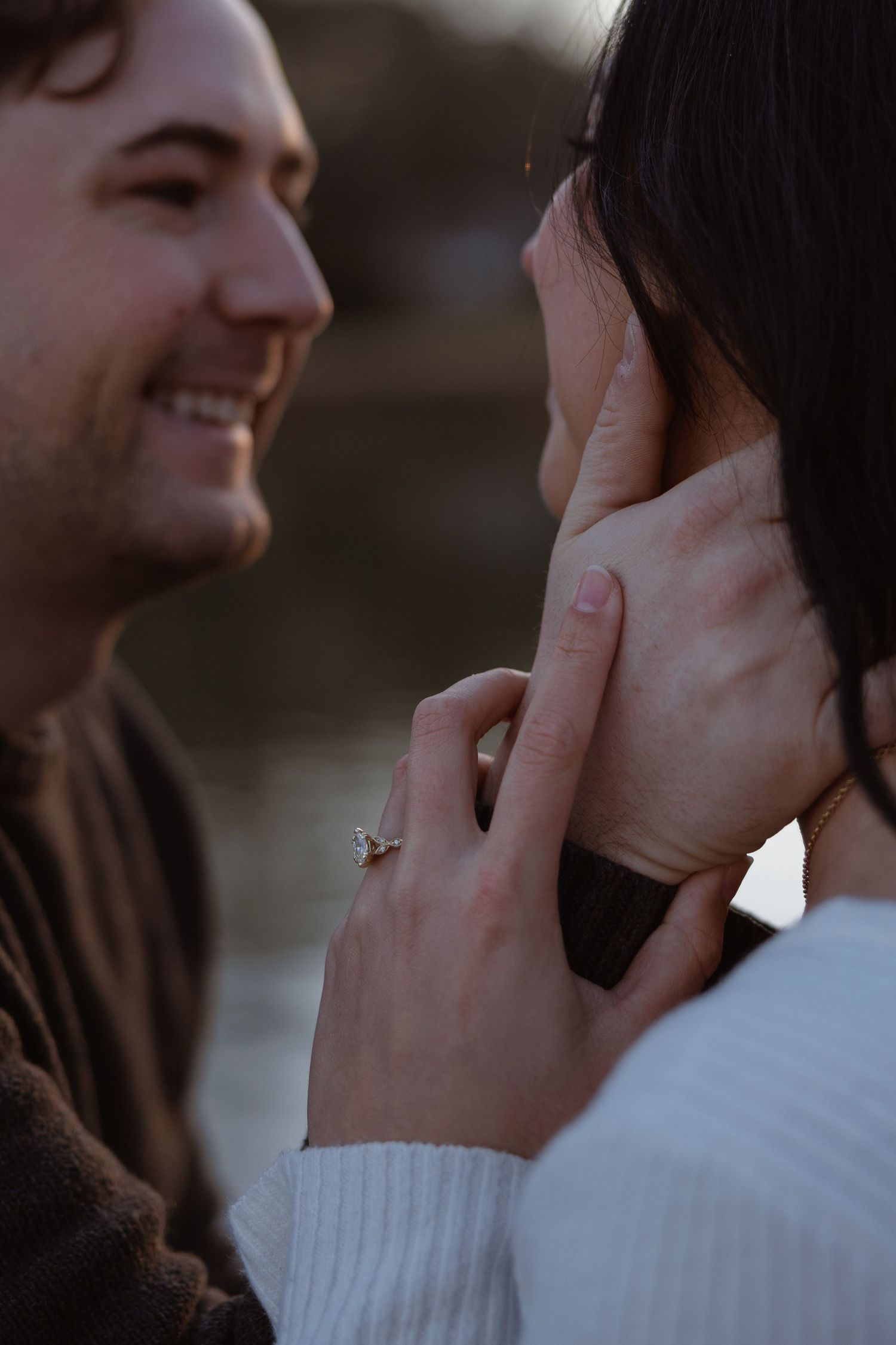 Couple sharing a tender moment, woman wearing a diamond engagement ring gently touching her partners face outdoors.