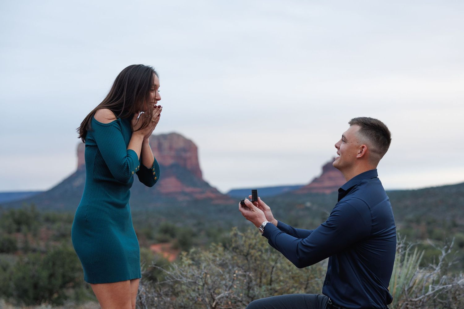 Man proposes to woman on one knee with ring box in Sedona Arizona red rock landscape at dusk.