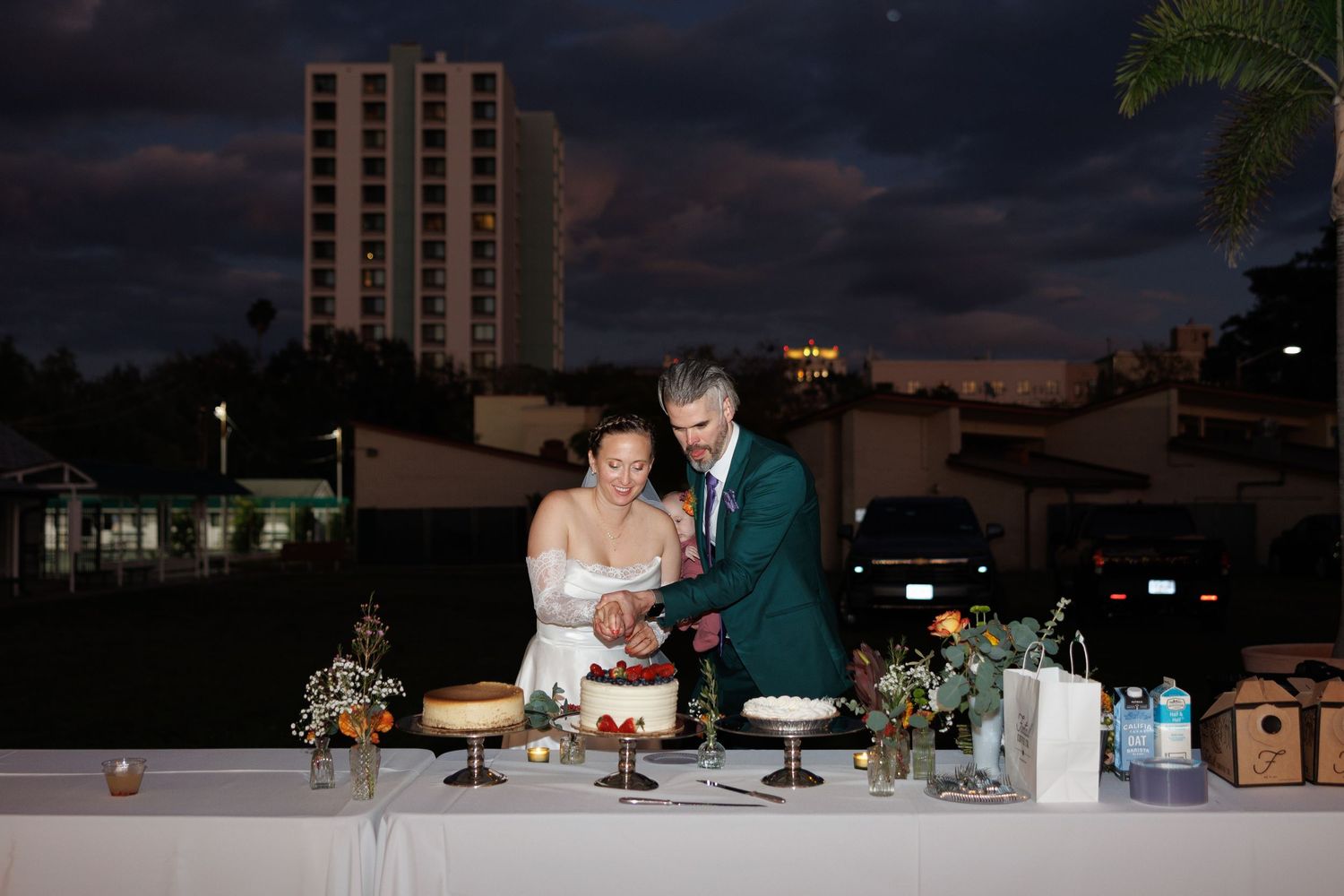 Bride and groom cutting wedding cake together outdoors at dusk with city lights behind.