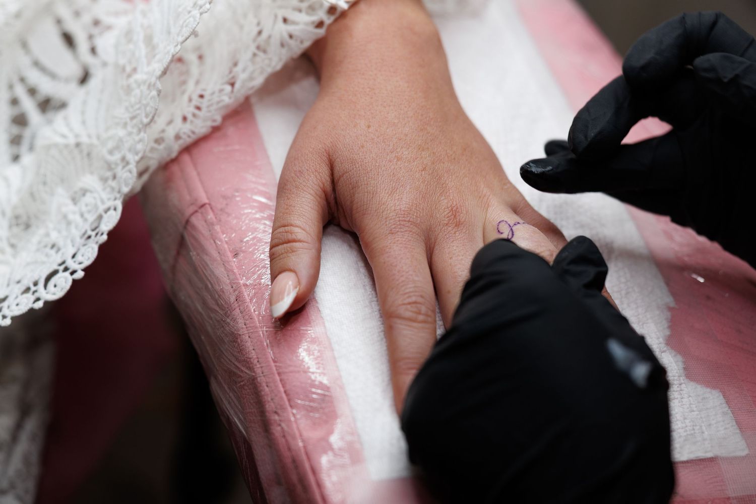 Tattoo artist in black gloves applying a small finger tattoo on a bride's hand.