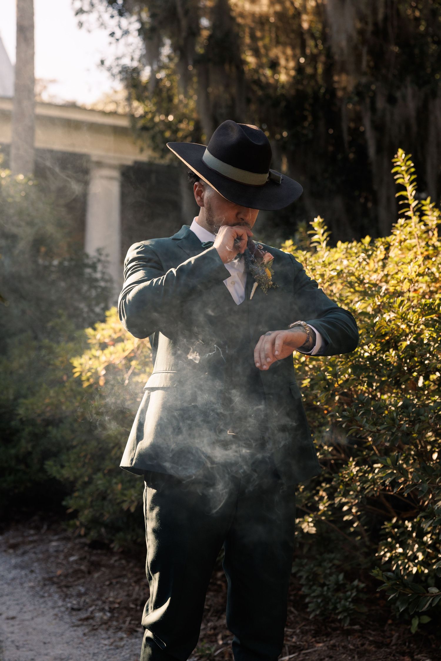 Groom in dark green suit and black cowboy hat checking his watch while smoking a cigar outdoors.