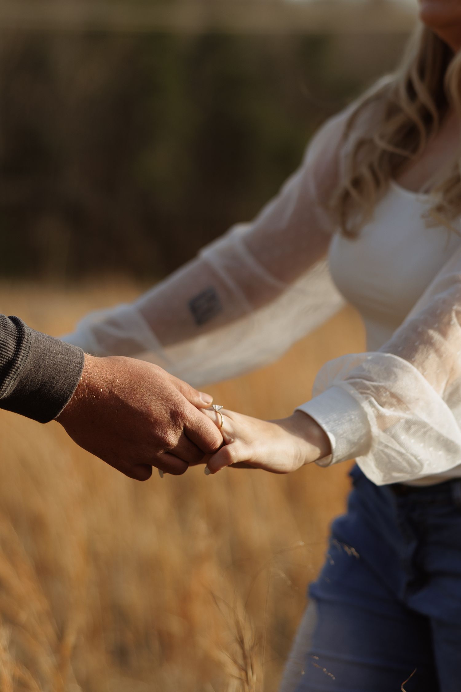 Couple holding hands in a golden wheat field, woman wearing white lace blouse and engagement ring.