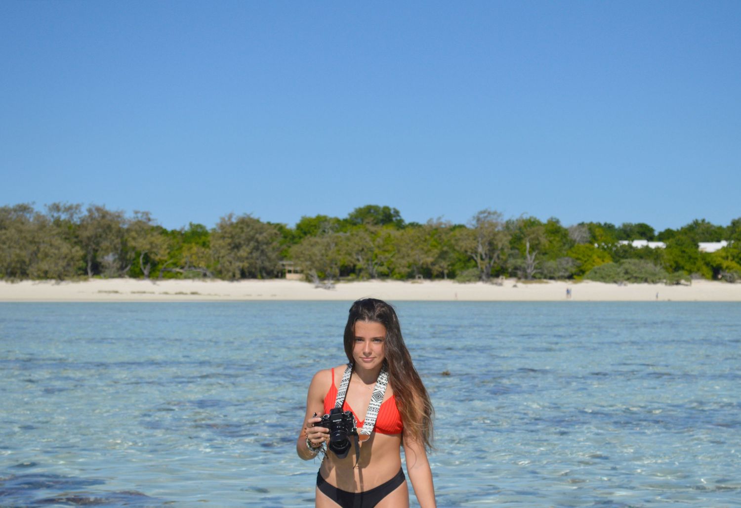 Woman in red bikini standing in shallow turquoise ocean water with sandy beach and green trees in background.