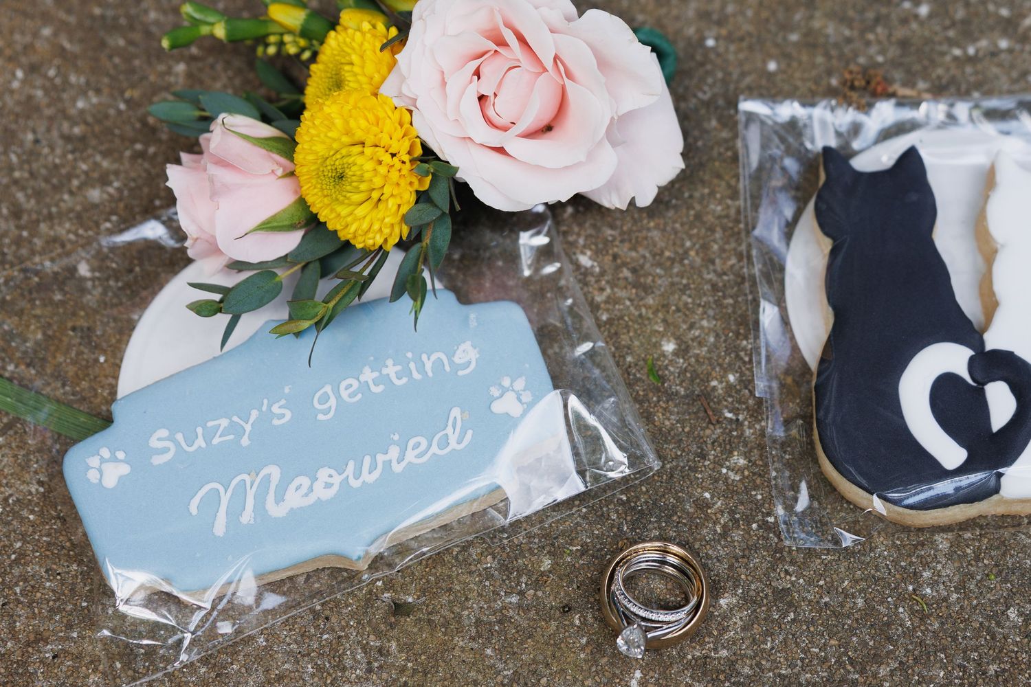 Wedding flat lay with cat-themed cookies, rings, floral bouquet, and a blue sign reading Suzys getting Meowied.