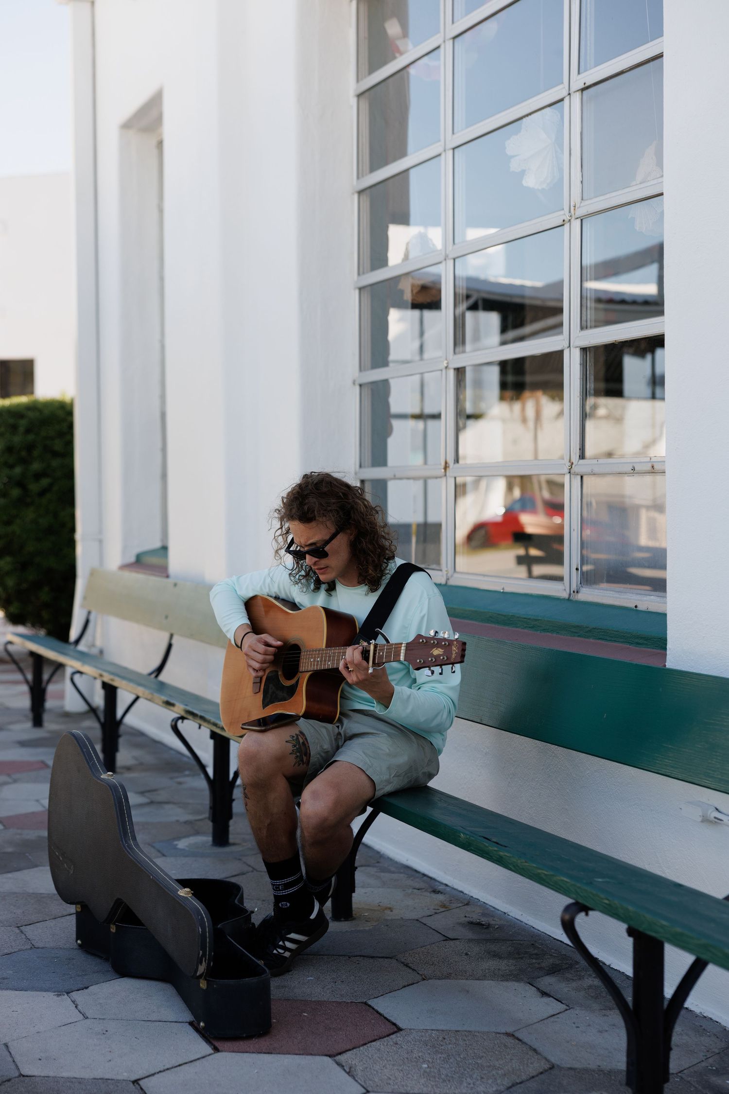 A curly-haired musician in sunglasses plays acoustic guitar on a green bench outside a white building.