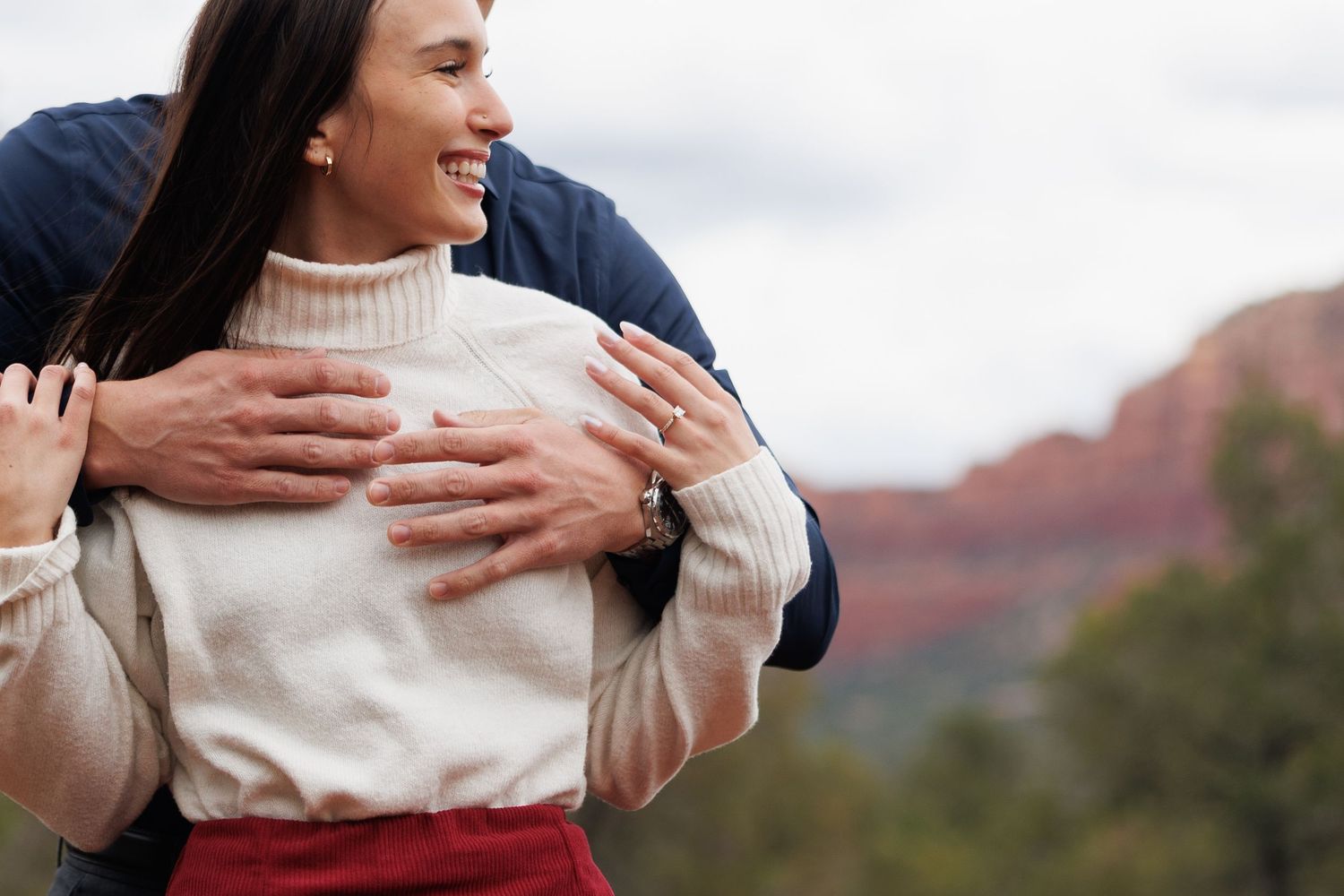 Man embracing woman from behind showing engagement ring with stunning Sedona red rock landscape in background.