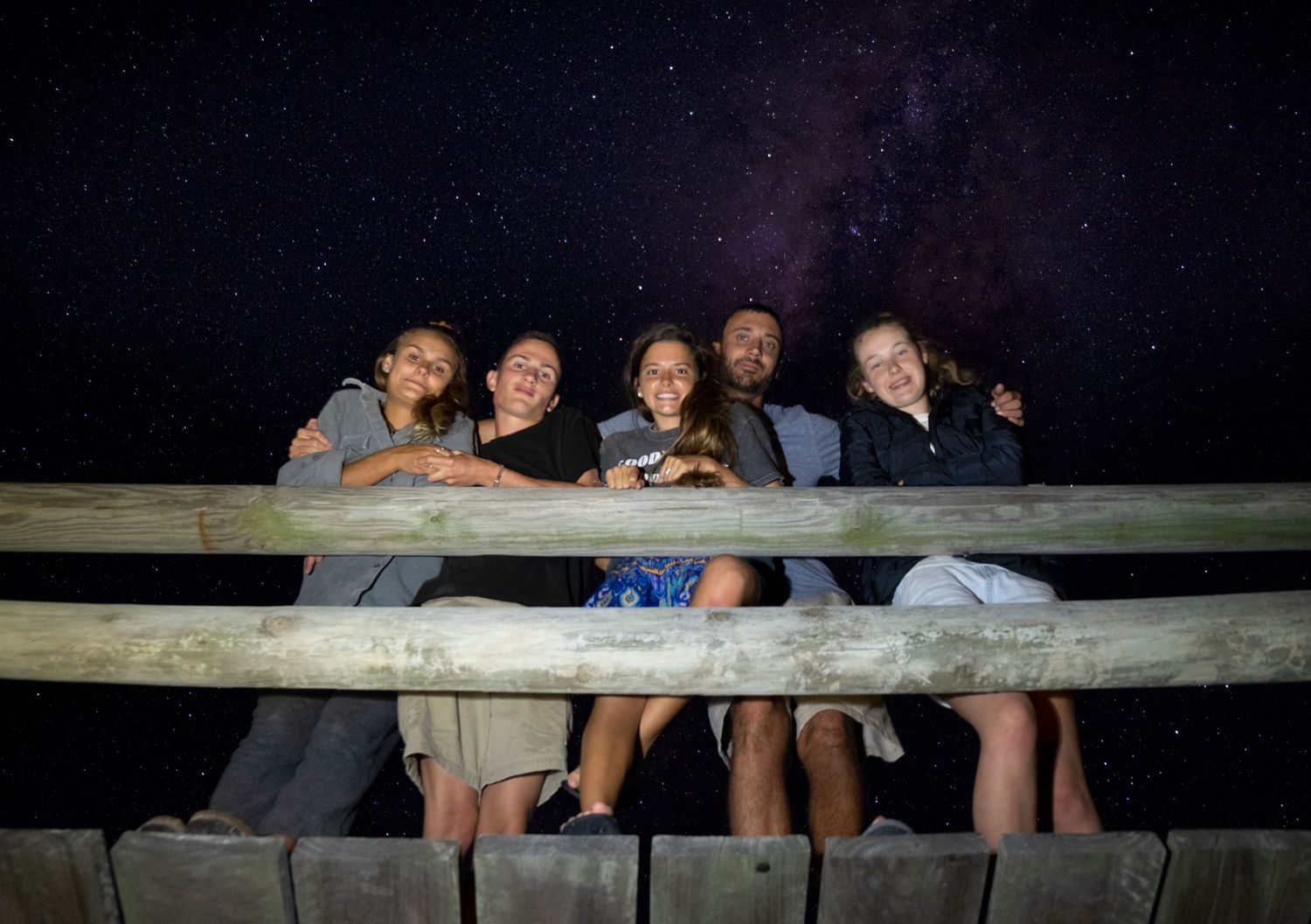 Group of friends posing together on a wooden fence under a stunning starry night sky.