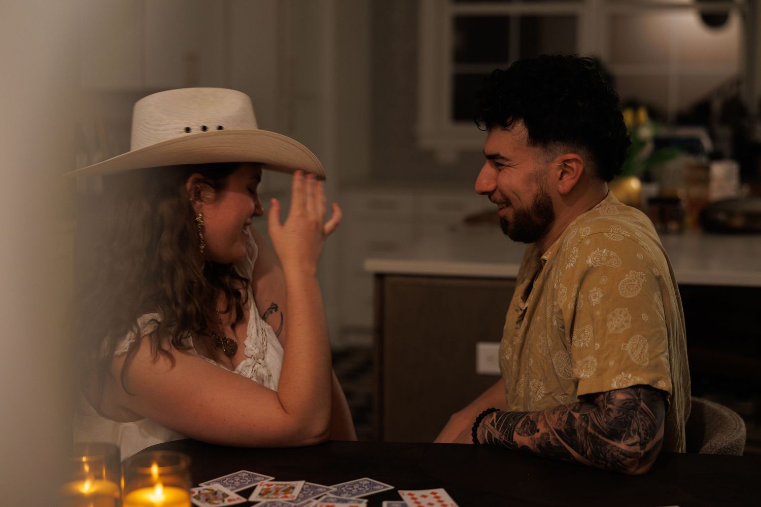 Woman in cowboy hat laughing with tattooed man by candlelight at table with tarot cards.