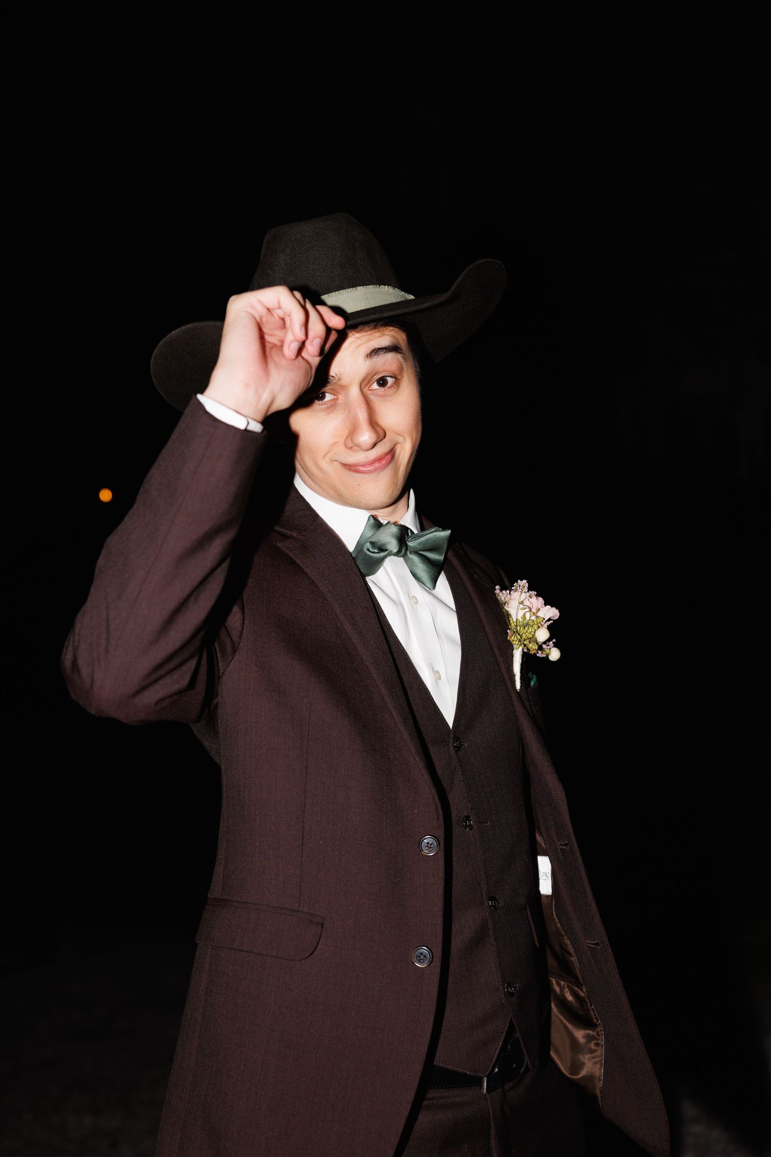Groom in maroon suit tips black cowboy hat while smiling against a dark night background.