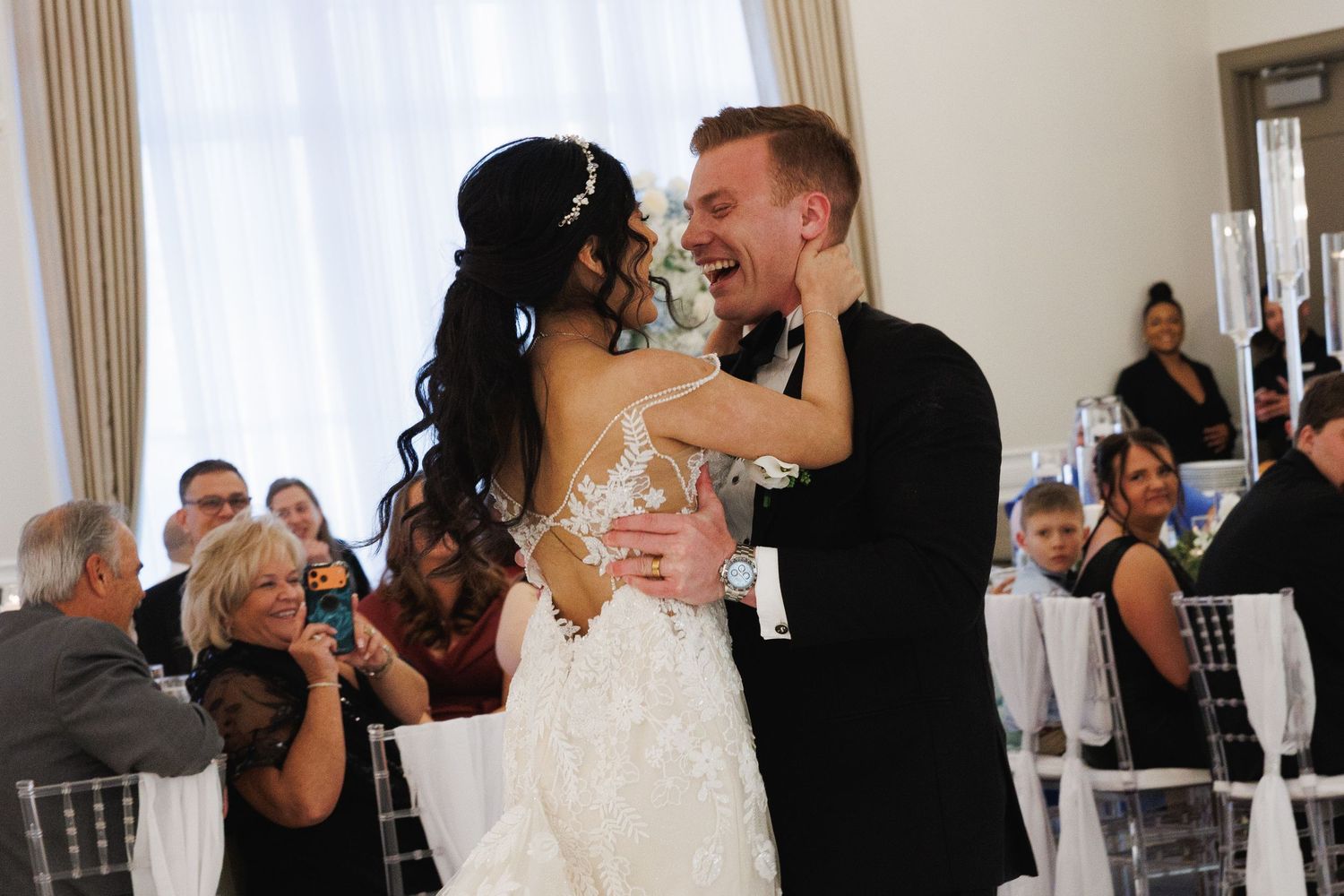 Bride and groom sharing their first dance, laughing joyfully in a decorated wedding reception hall.
