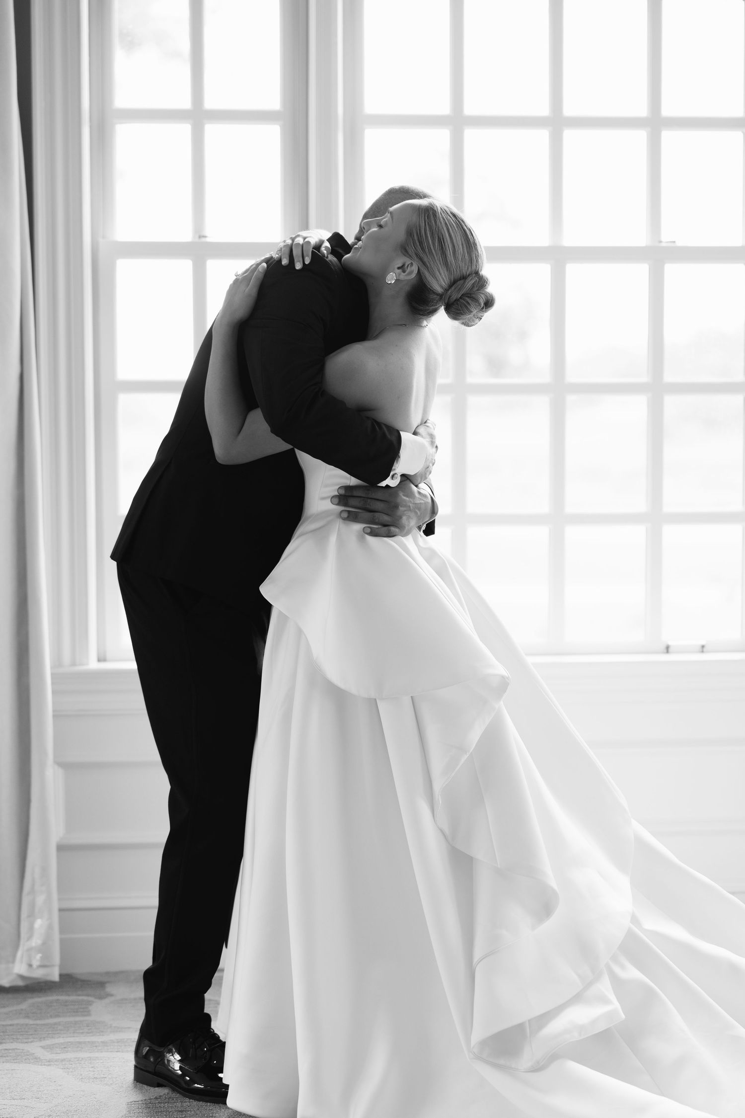 Black and white photo of bride and groom sharing a tender embrace near a large window on their wedding day.