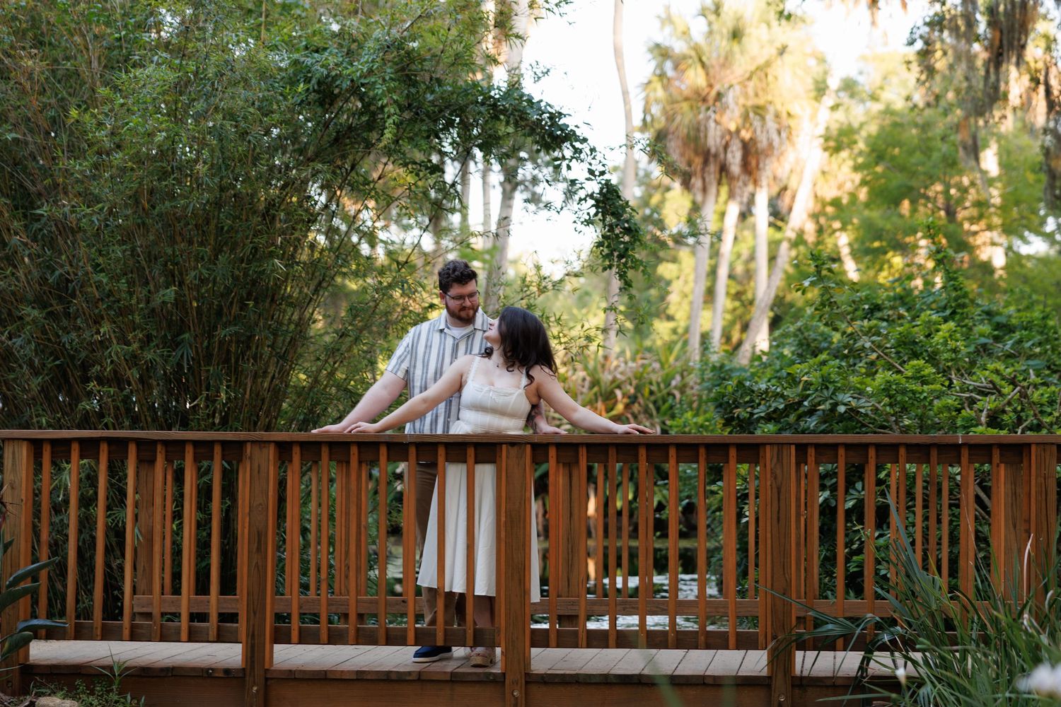 Couple standing on a wooden bridge surrounded by lush green trees and palm trees during golden hour.