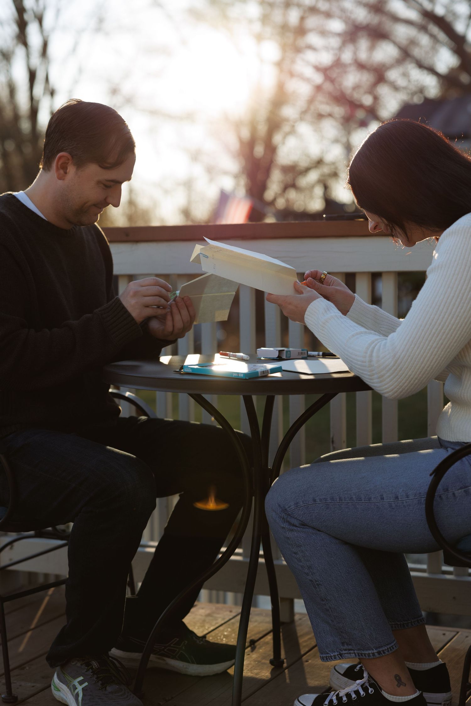 Man and woman reviewing paper airplanes at an outdoor table during a romantic golden hour sunset.