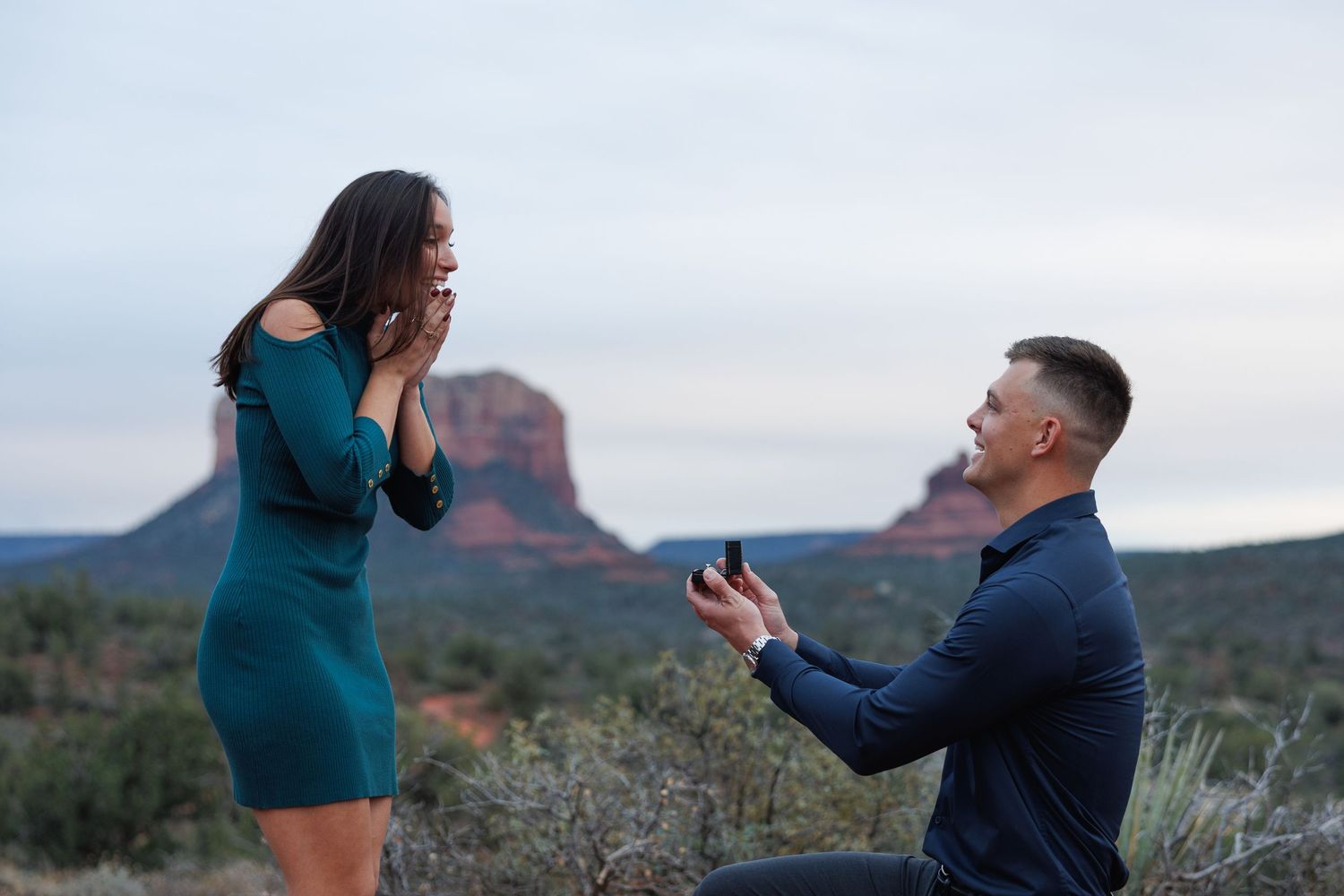 Man proposes to woman on one knee in Sedona Arizona with red rock formations in the background.