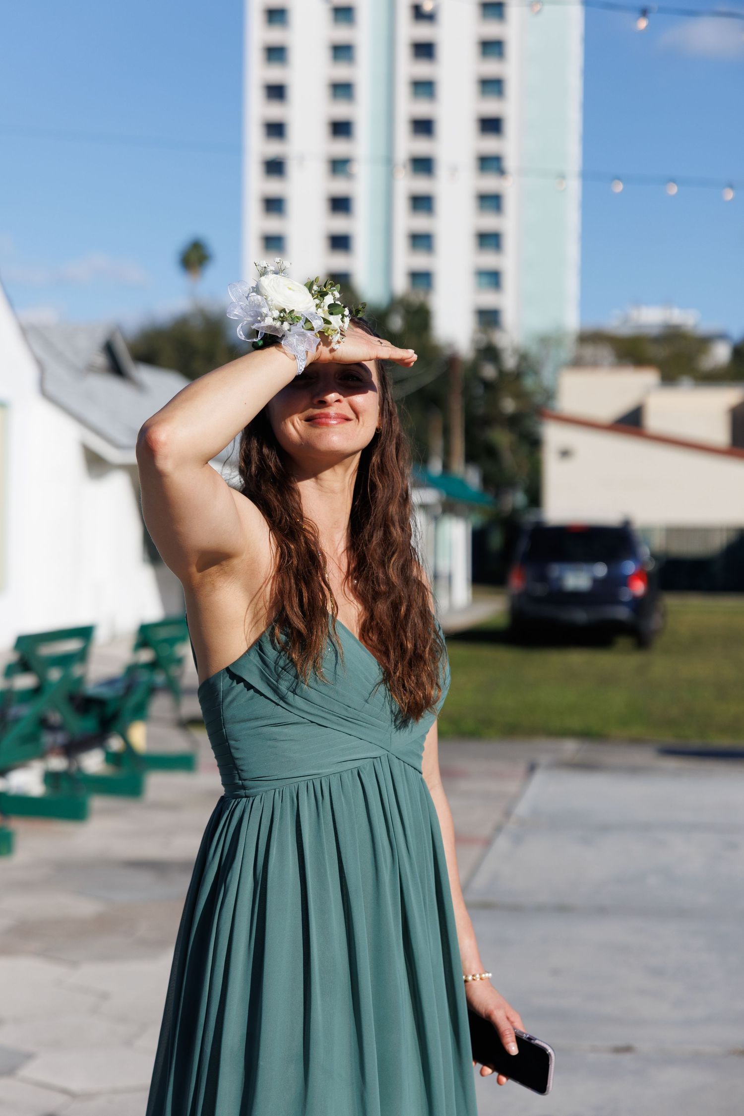 Woman in teal bridesmaid dress shields eyes from sun while holding flowers at outdoor wedding venue.