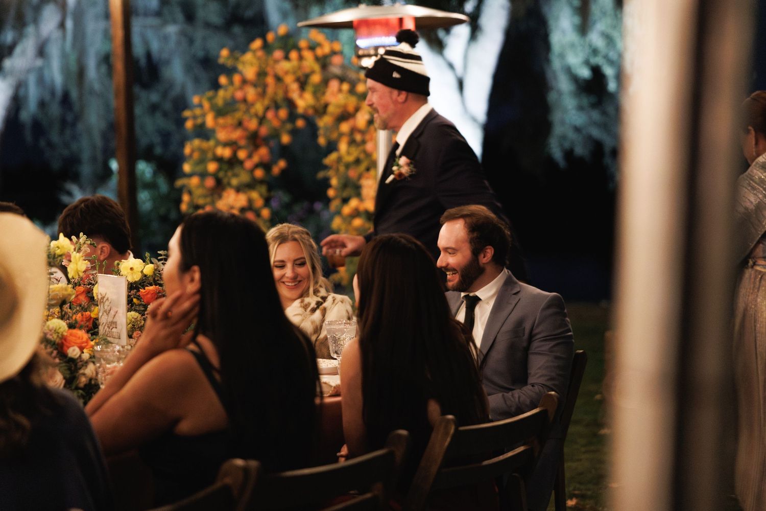 Wedding guests laugh and chat at elegantly decorated outdoor reception table under warm lighting.