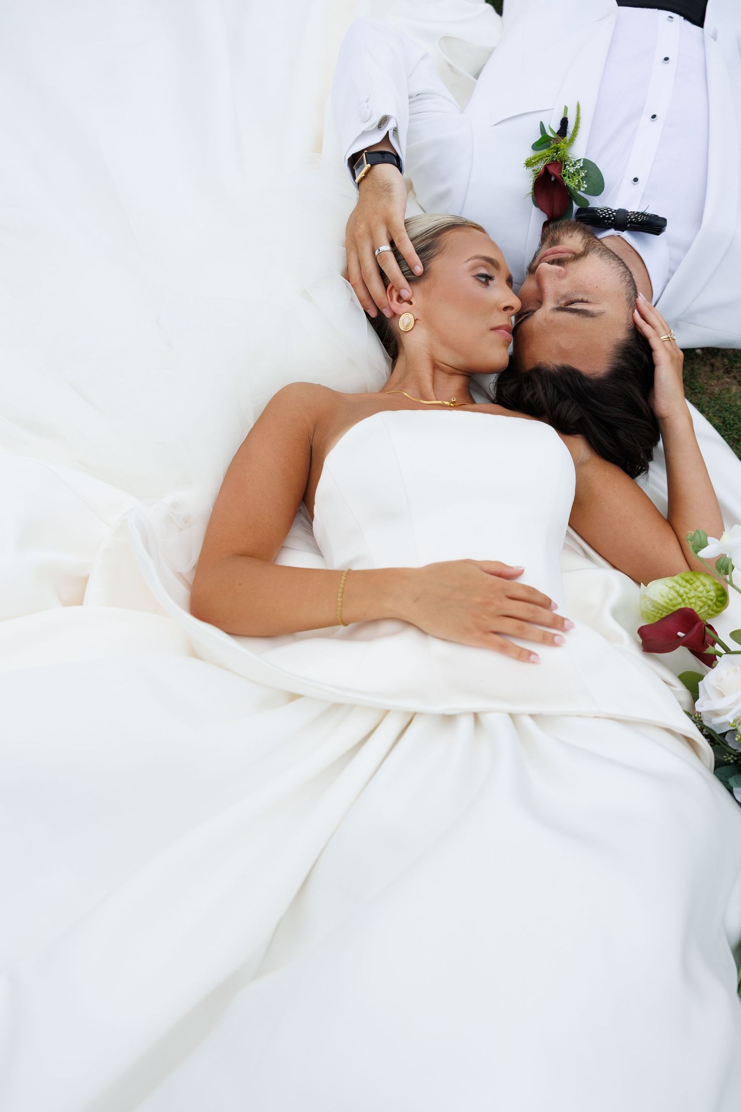Bride and groom lying on white wedding dress fabric sharing a romantic kiss from an aerial perspective.