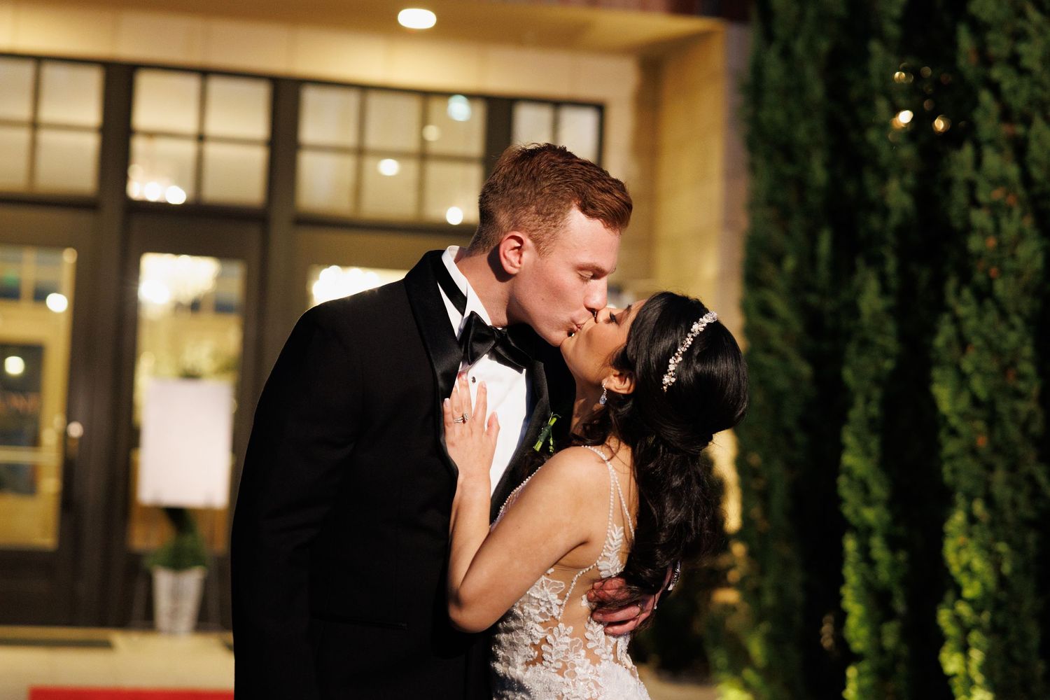 Bride and groom sharing a romantic kiss outdoors at night in front of an elegant venue.